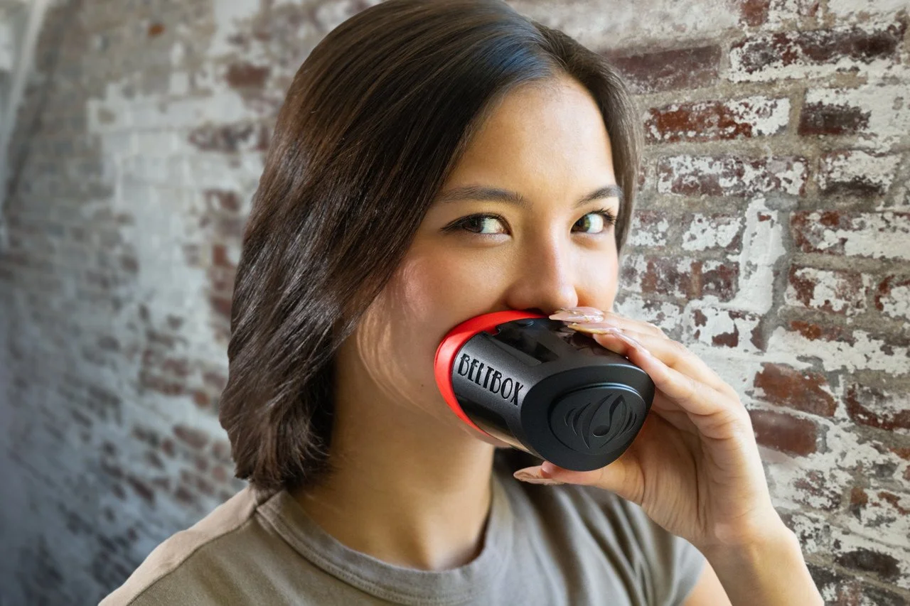 A woman with short brown hair holding a black and red coffee mug with the word 'BELIBOX' on it, standing in front of a brick wall.