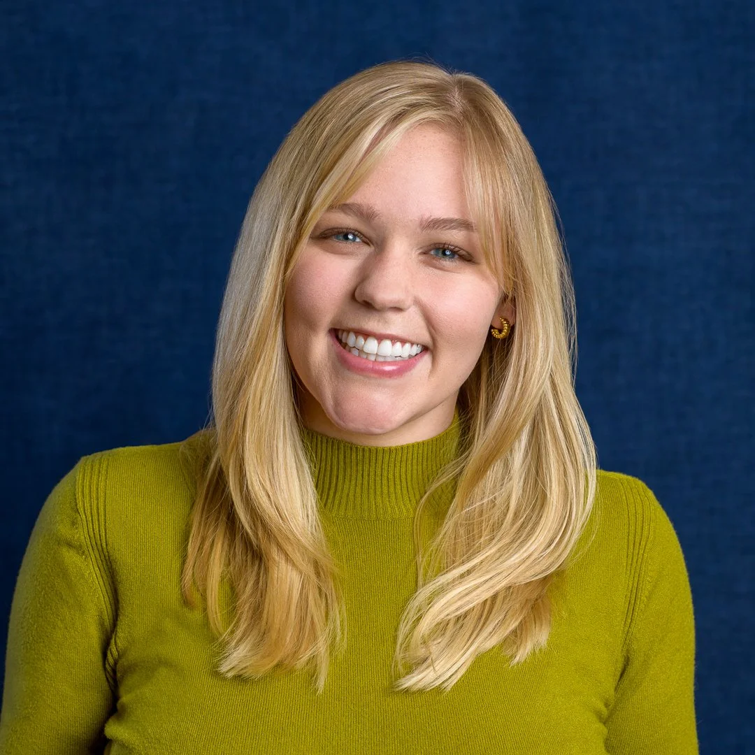 A young woman with long blonde hair, blue eyes, and a bright smile, wearing a green turtleneck sweater and gold hoop earrings, standing against a dark blue background.