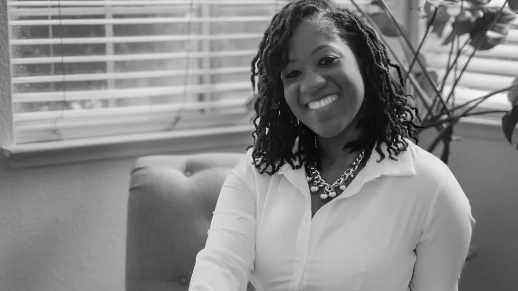 A smiling woman with curly hair sitting near a window with blinds, wearing a white shirt and a chunky necklace.