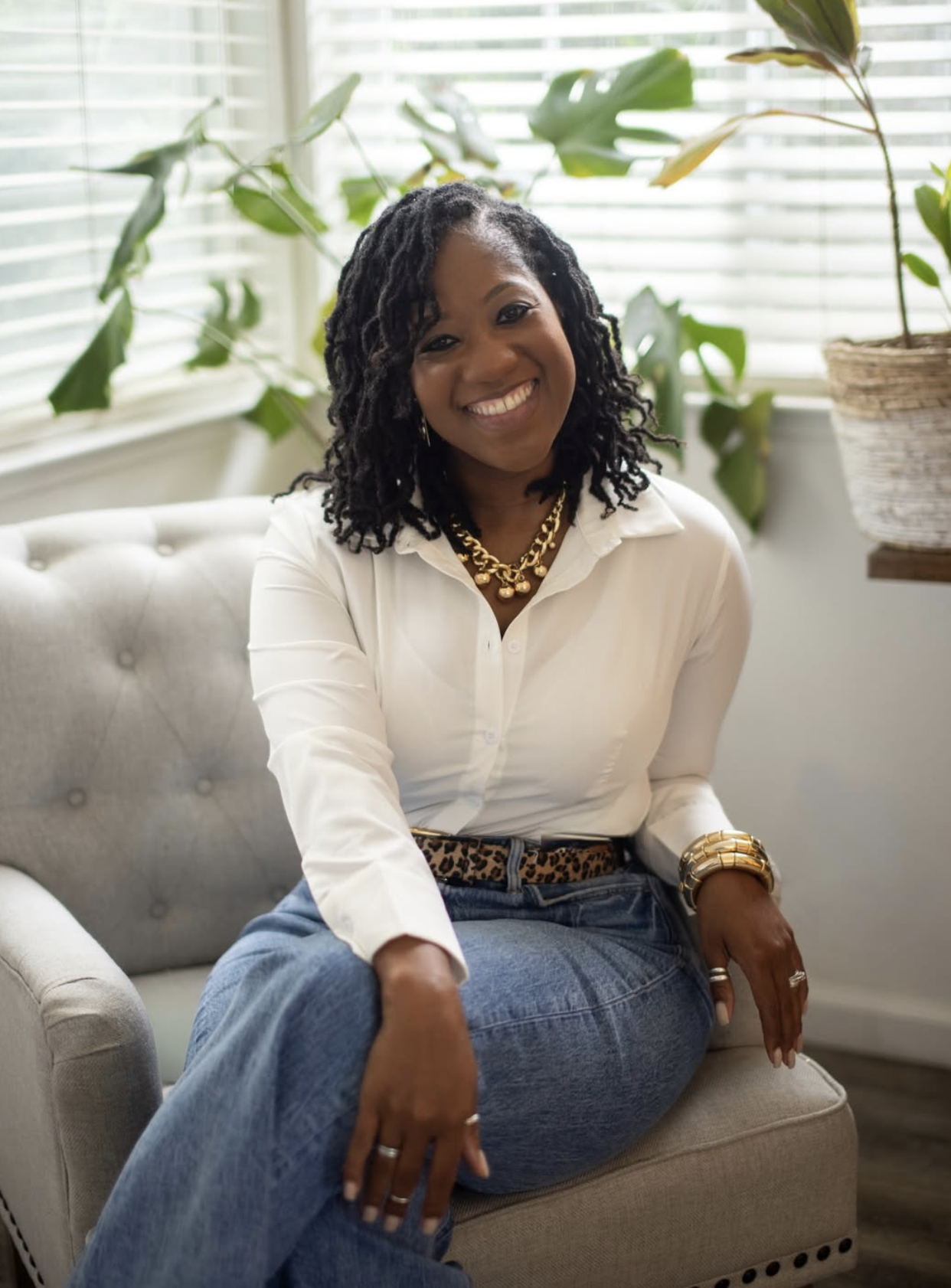 A woman with black hair styled in twists, smiling, sitting on a light gray sofa in a room with large window blinds and green plants.