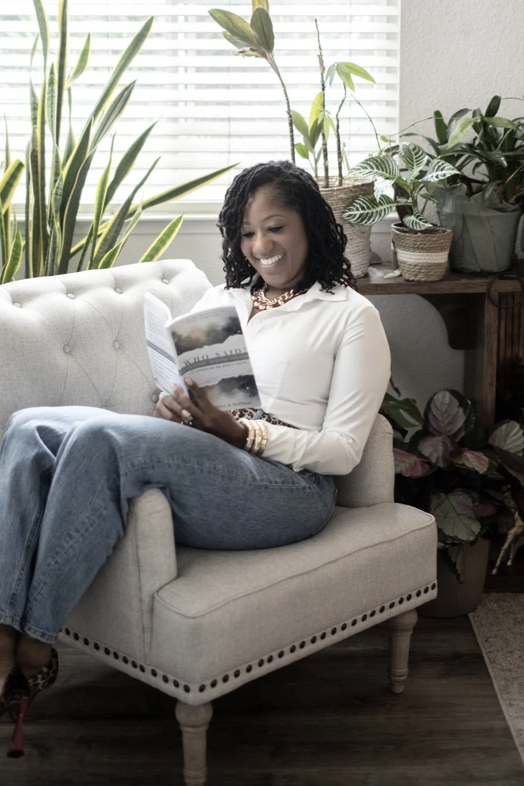 A woman sitting on a light-colored sofa, reading a book or magazine, surrounded by green plants near a window with blinds.
