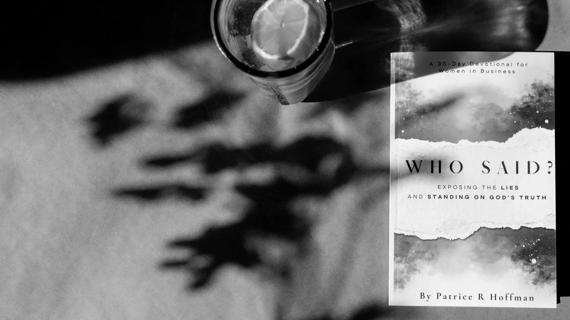 A black and white photo of a book titled "Who Said?" by Patrice R Hoffman, placed on a surface along with a glass of water with lemon slices, with the book’s cover featuring an abstract mountain landscape and space imagery.