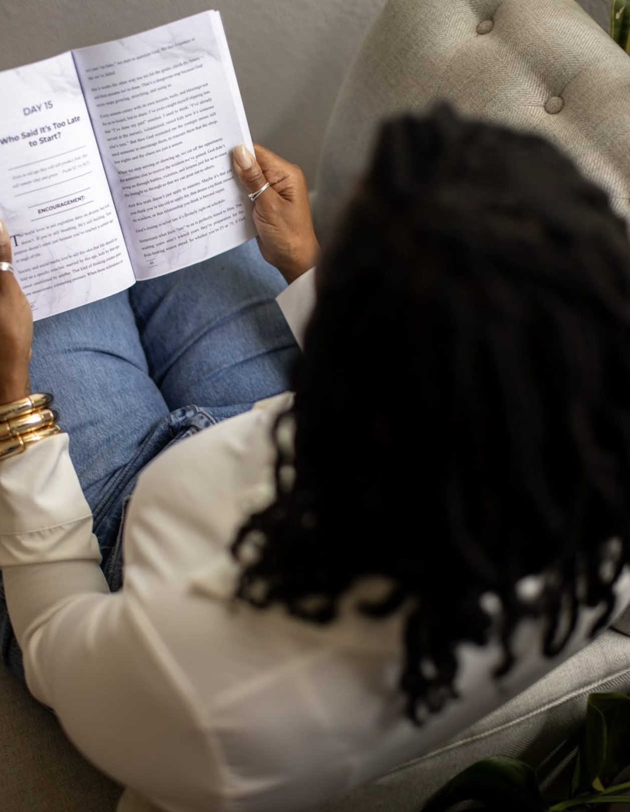 Person sitting on a beige chair, reading a booklet, wearing a white shirt, blue jeans, multiple gold bracelets, and rings.