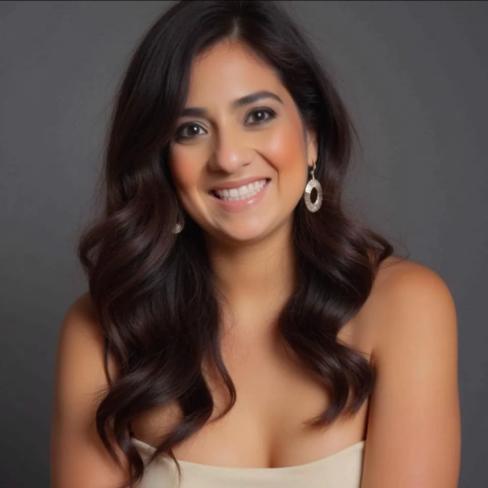 A woman with long, dark wavy hair smiling at the camera, wearing earrings and a cream-colored off-shoulder top against a gray background.