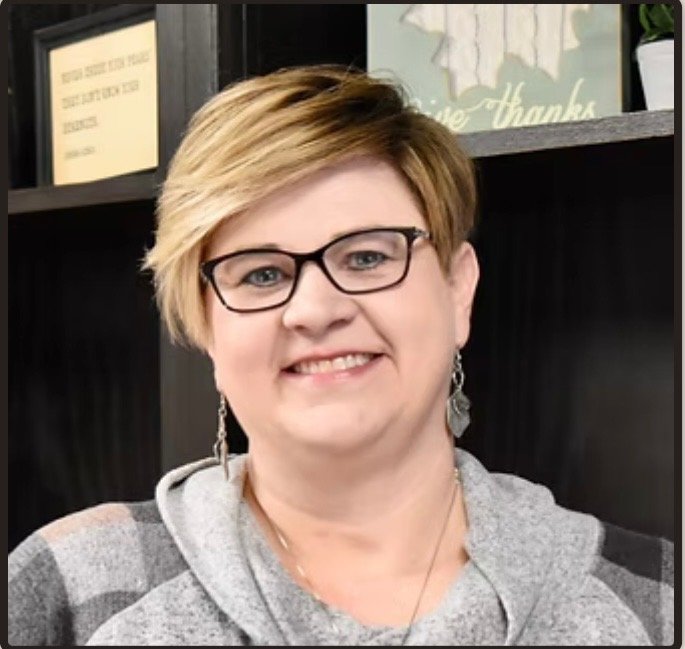 Smiling woman with short blonde hair, glasses, and earrings, wearing a gray hoodie, standing indoors with dark shelves and a sign that says 'Thanks' in the background.