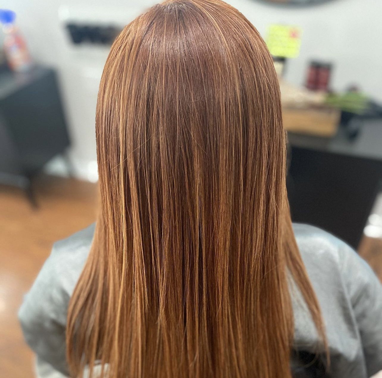 Back view of a woman with long, straight, shiny reddish-brown hair in a salon.