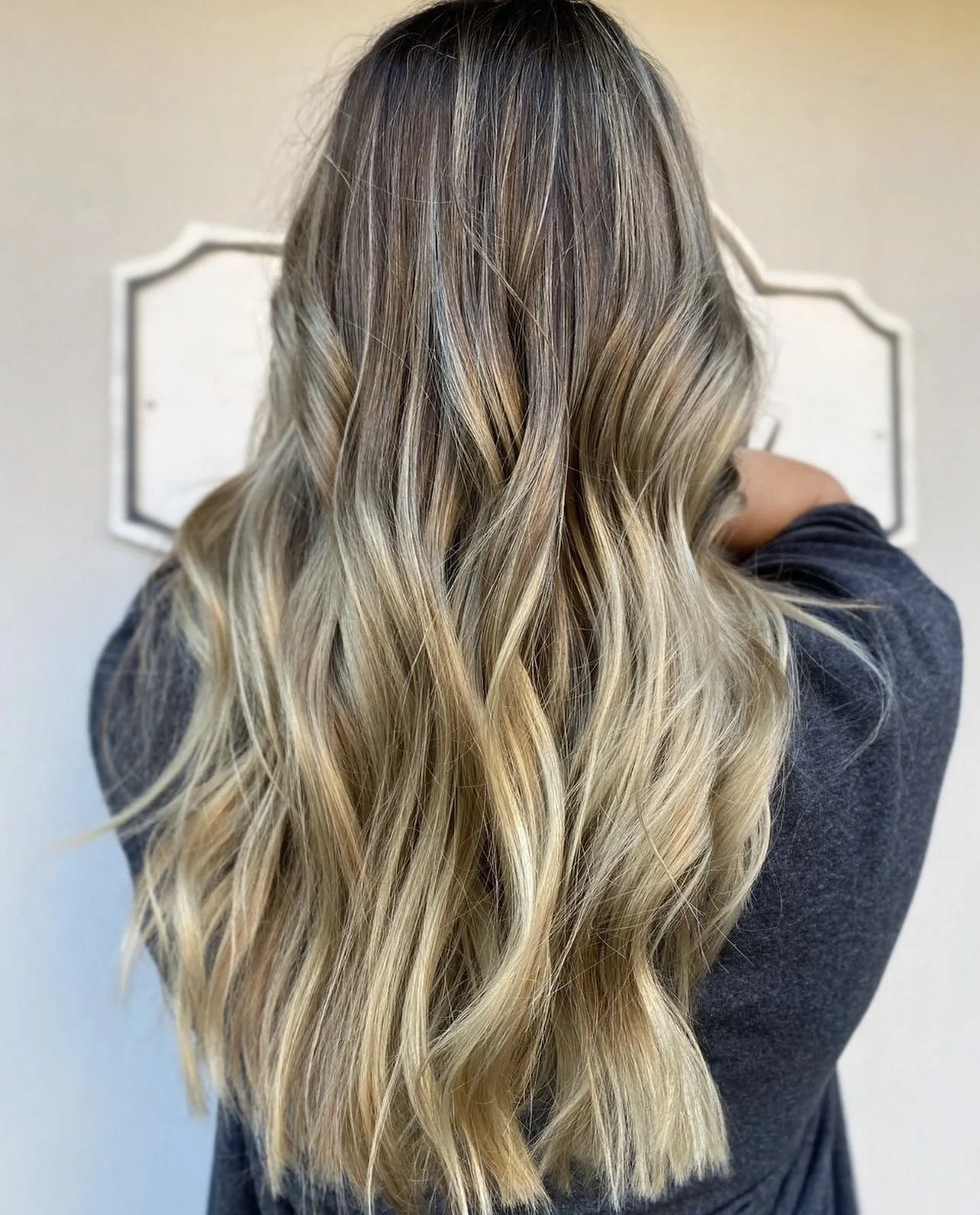 Back view of woman with long, wavy blonde hair, wearing a dark gray top and standing in front of a white wall with a decorative mirror.