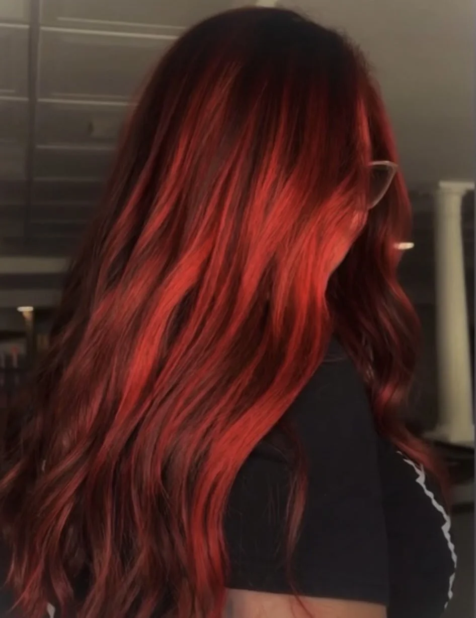 A woman with long, wavy hair dyed in shades of red and black, wearing glasses, in an indoor setting.