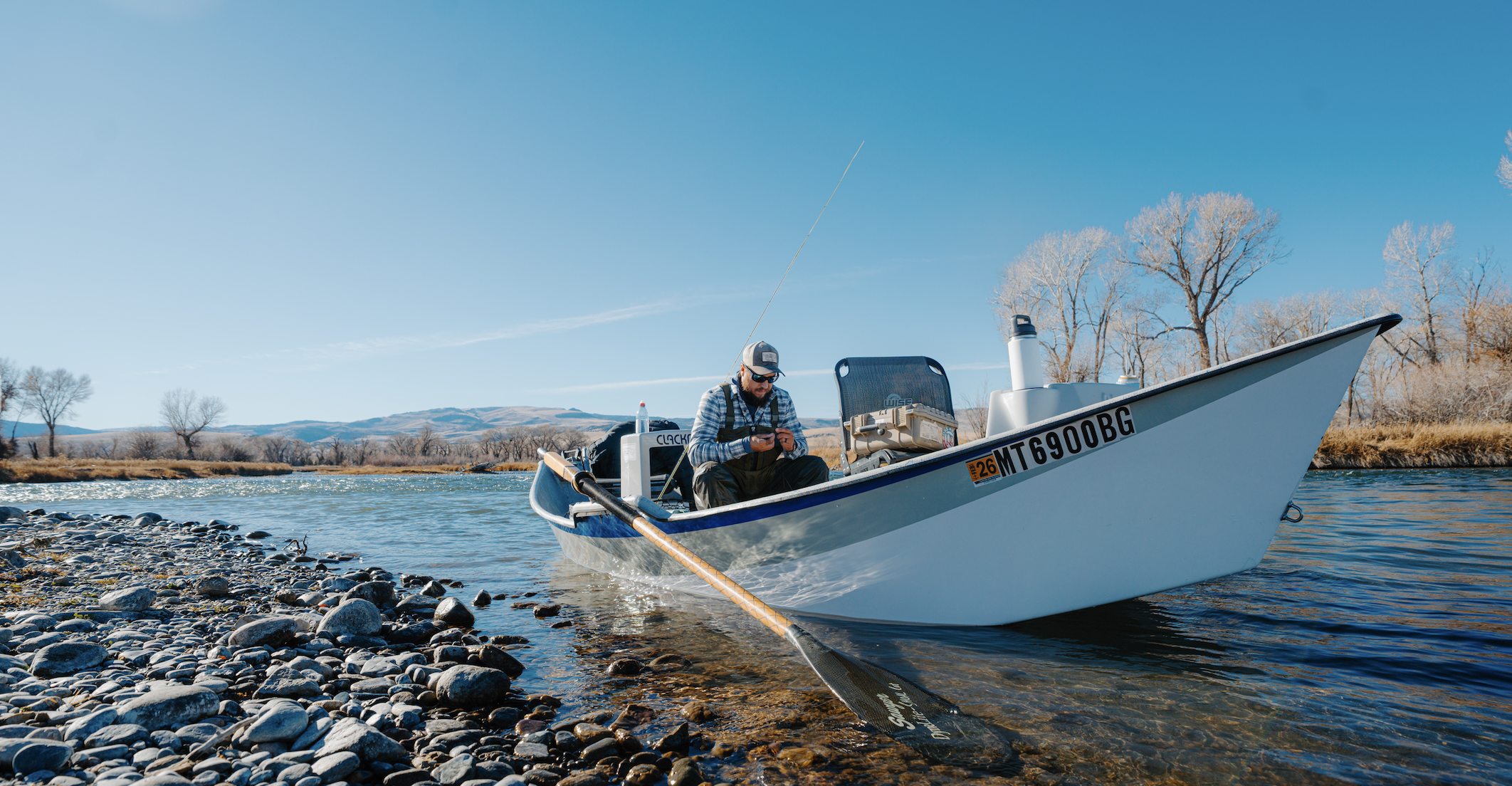 A man sitting in a drift boat on the madison river, looking at his fly fishing flies. The boat is on a rocky shore with a clear blue sky and leafless trees in the background.