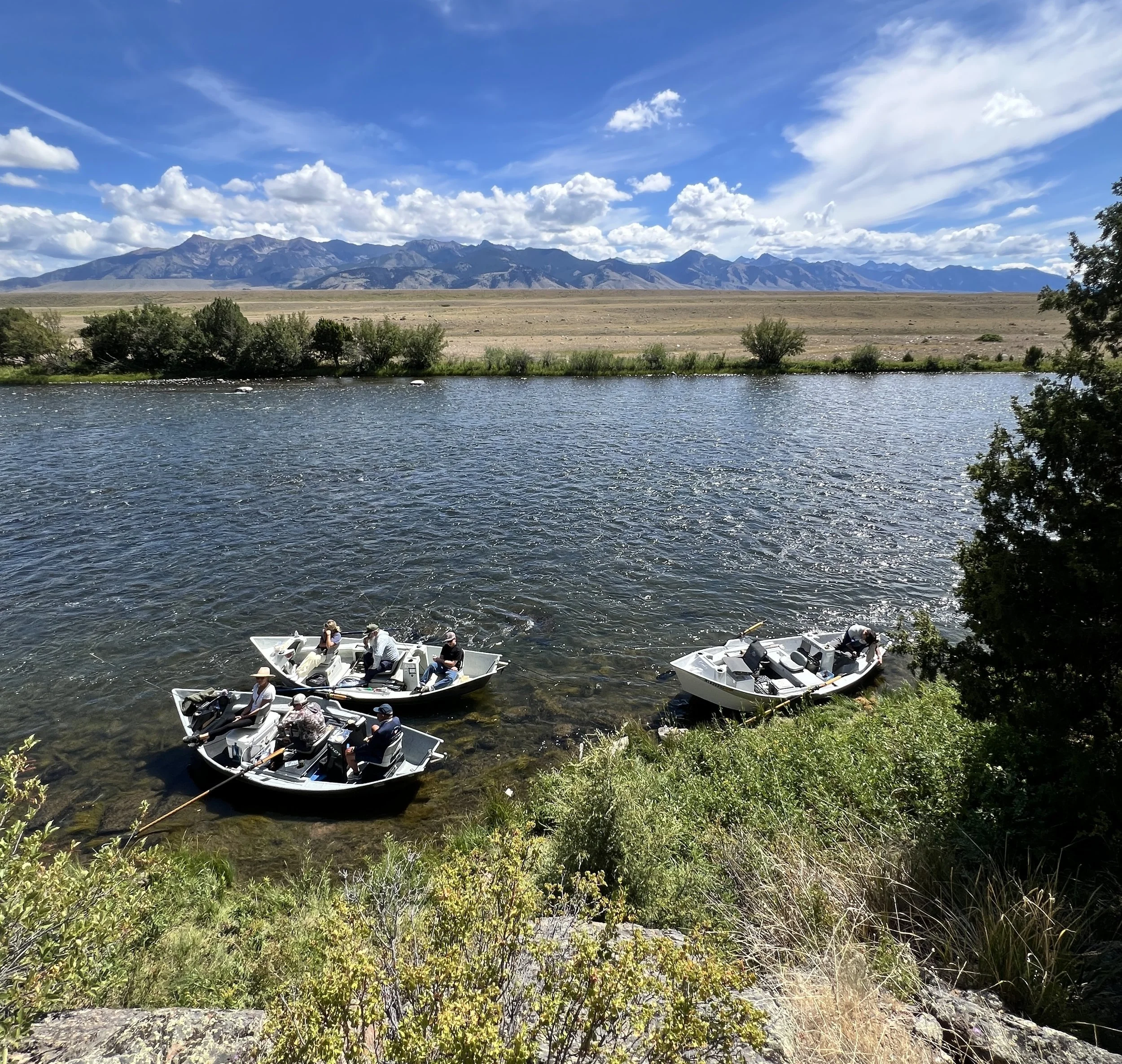 People fishing from boats on the Upper Madison River with mountains in the background and a partly cloudy sky. They are on a Group Guide Trip with Rising Trout Fly Fishing Outfitters in Bozeman