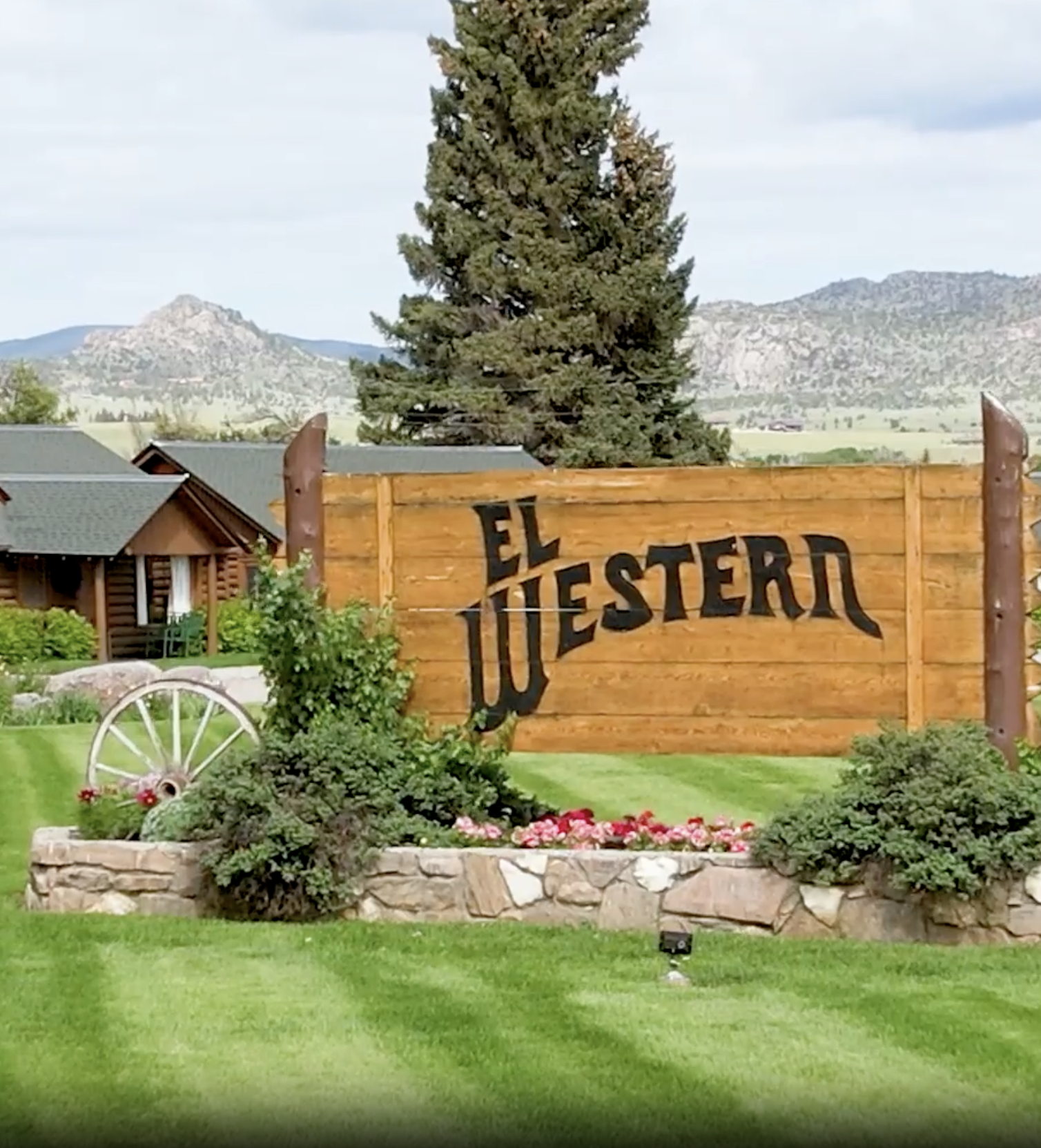 Sign for El Western restaurant in a landscaped outdoor area with grass, flowers, bushes, a stone border, a wooden wagon wheel, and a large tree in the background.