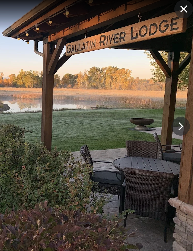 A covered outdoor patio area overlooking a pond with trees in the background. The patio has a wooden sign that says 'Gallatin River Lodge' and features wicker chairs and a round table.