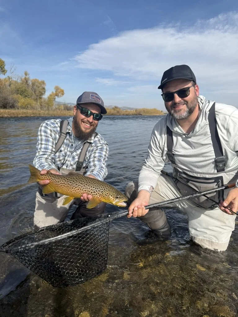 Two men in outdoor gear kneeling in the madison river, holding a large brown trout with yellow spots.