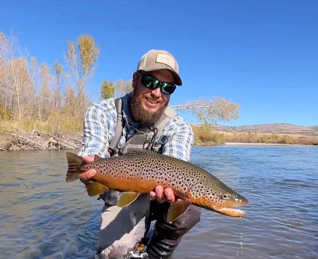 Man wearing sunglasses, a hat, and fishing gear holding a large brown trout in a river with fall trees and hills in the background.