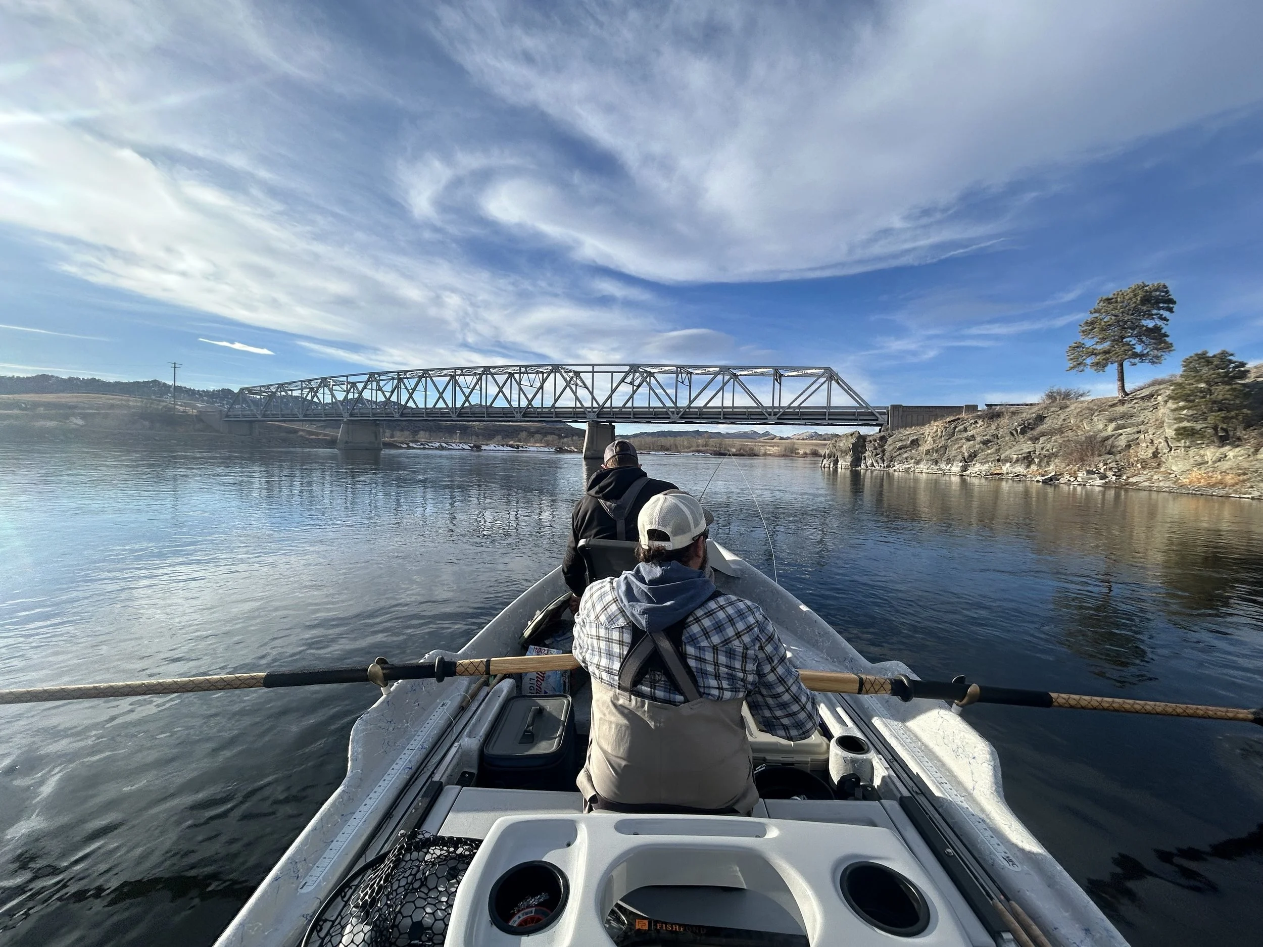 People fishing from a boat on the missouri river with the wolf creek bridge and trees in the background on a clear day.