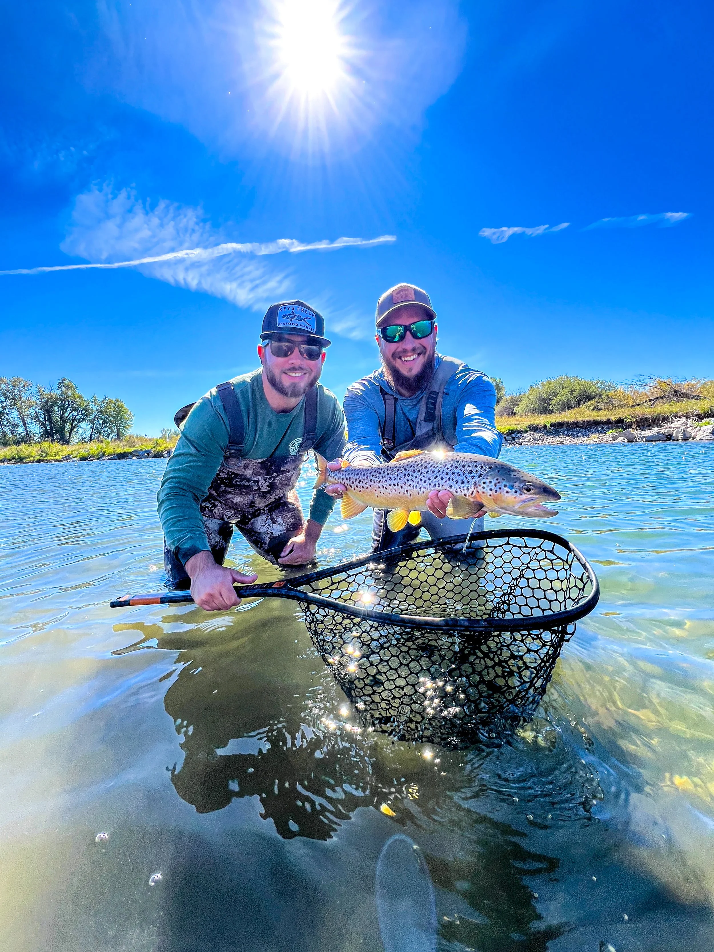 A fly fishing guide and client on the gallatin river, holding a large fish with a net. The sky is bright blue with sun rays shining directly overhead. Trees are visible in the background.