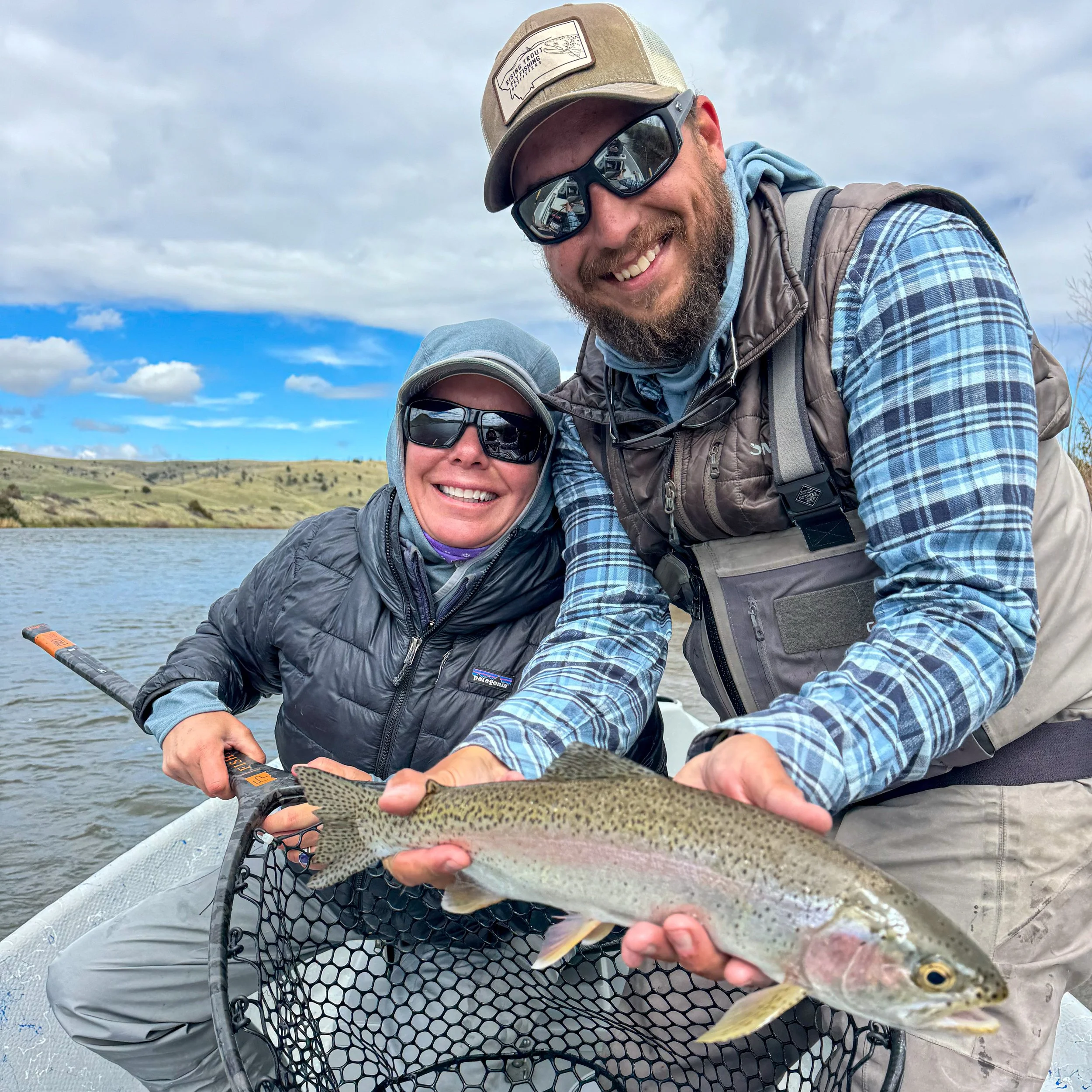 Two smiling people, a man and a woman, on a boat holding a large rainbow trout they caught, with a lake and hills in the background under a partly cloudy sky.