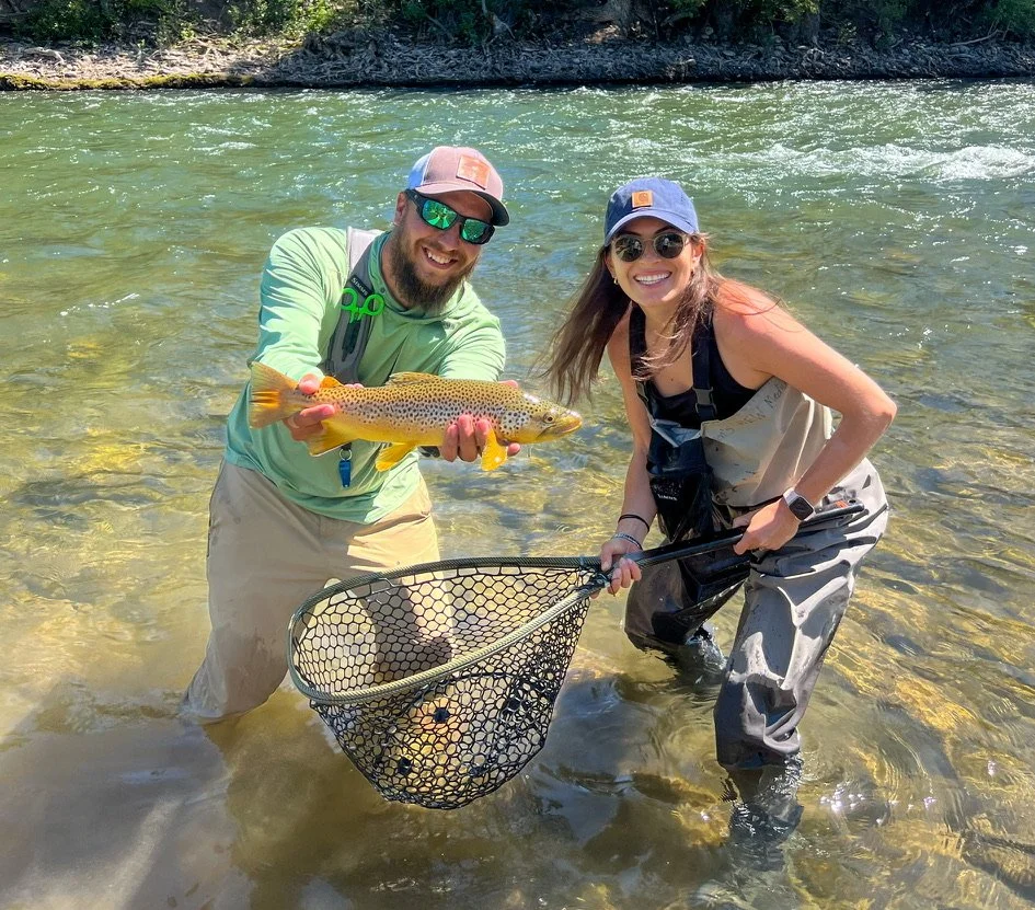 Two smiling people in fishing gear standing in shallow river water, holding a large fish with colorful scales, and a fishing net.