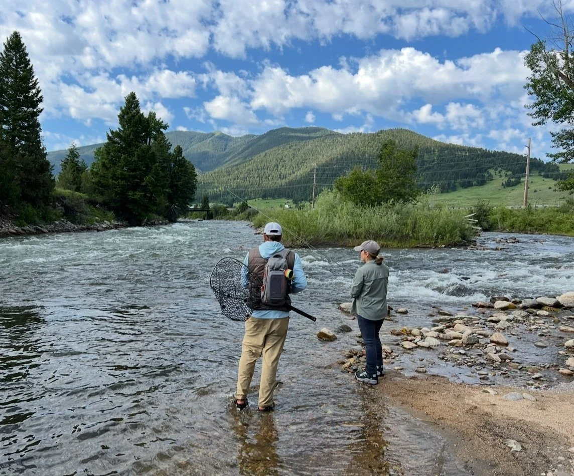 Two people standing in a river with fishing gear during a guided fly fishing walk wade trip, surrounded by trees, mountains, and a partly cloudy sky.