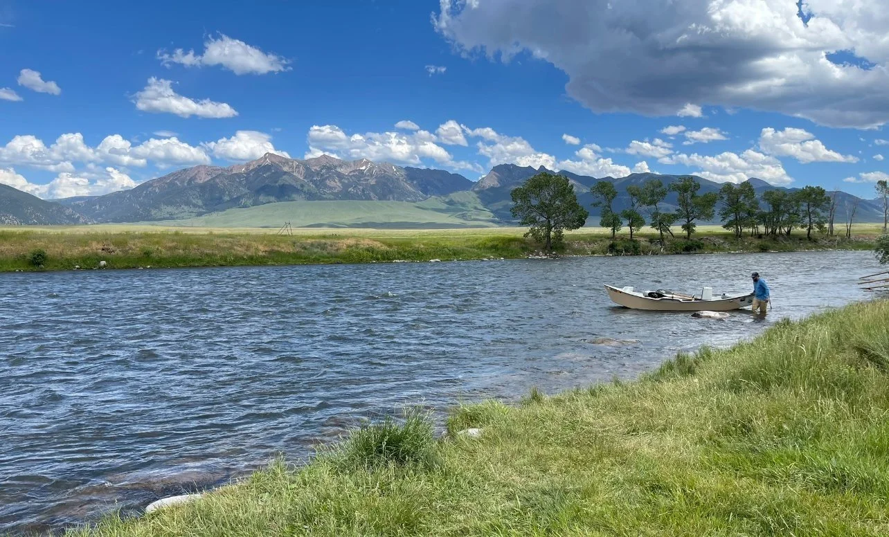 A person launching a drift boat into the upper madison river with a backdrop of green fields and mountains under a partly cloudy sky.