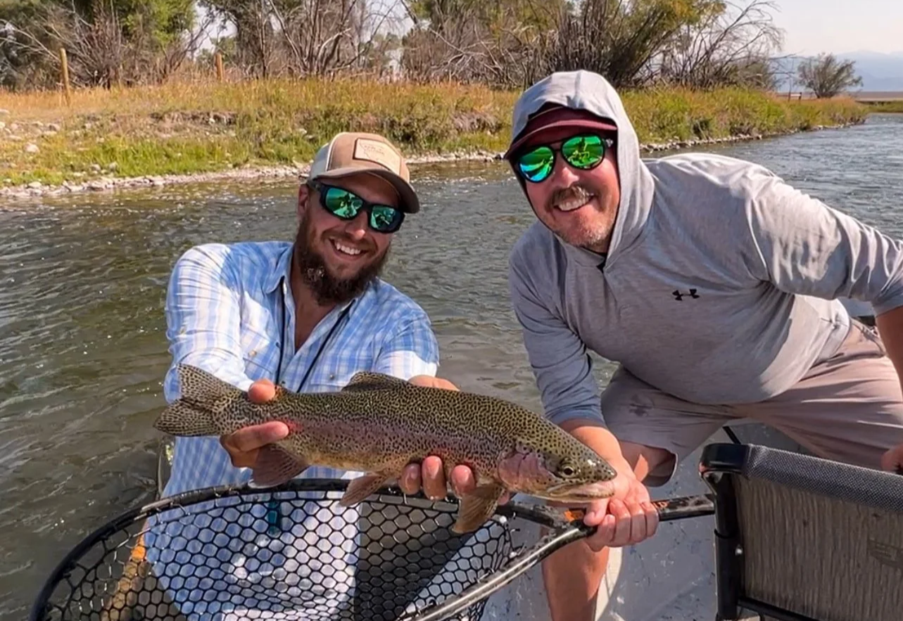 Two men smiling and holding a large rainbow trout they caught while fishing the Madison River, with trees and clear sky in the background.