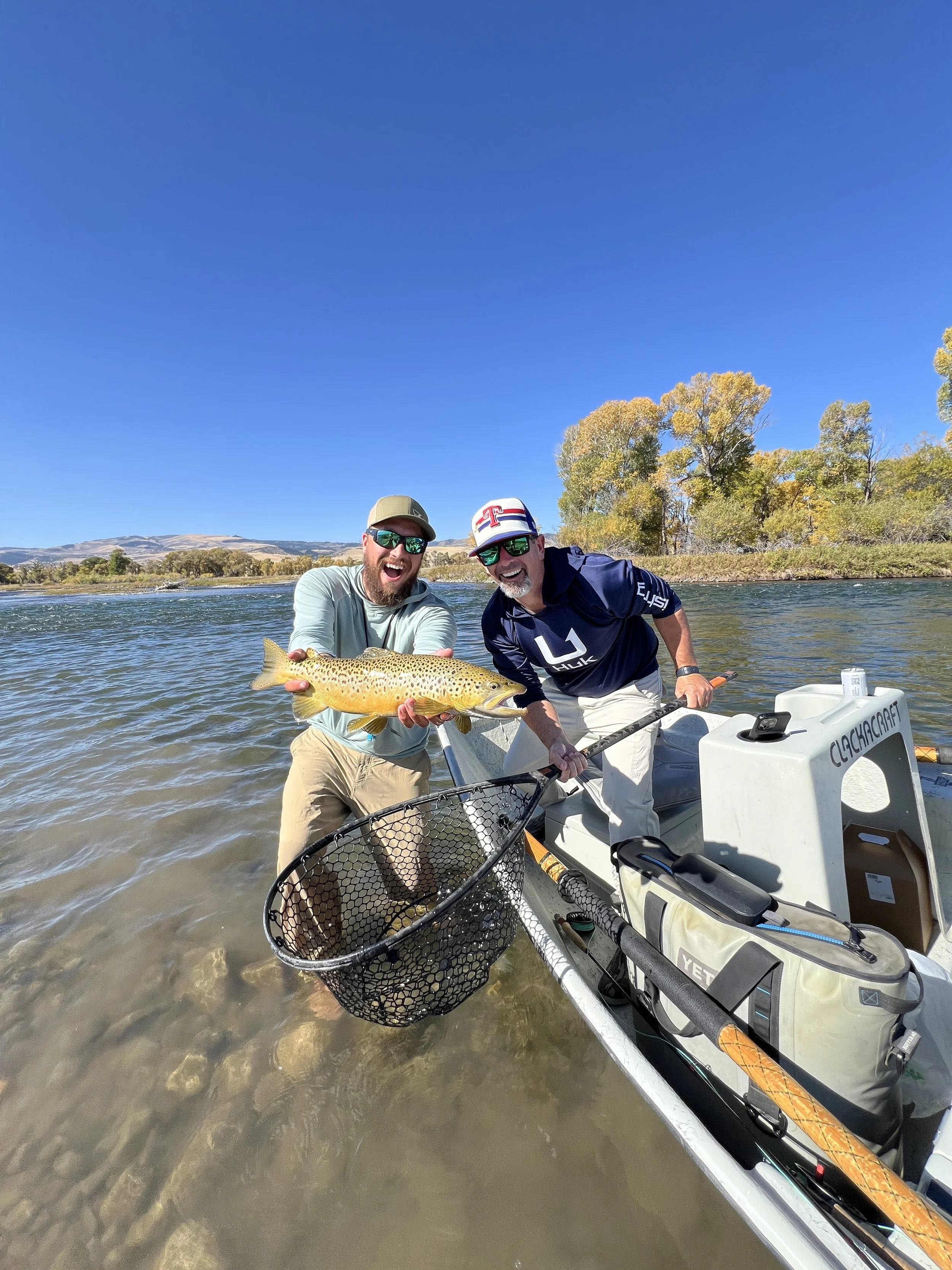 Two men standing in shallow water on a boat, holding a large fish, with clear blue sky and trees with autumn leaves in the background.