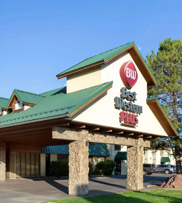 Exterior of a Best Western Plus hotel with a stone and stucco facade, green metal roof, and a large circular sign with the BW logo.