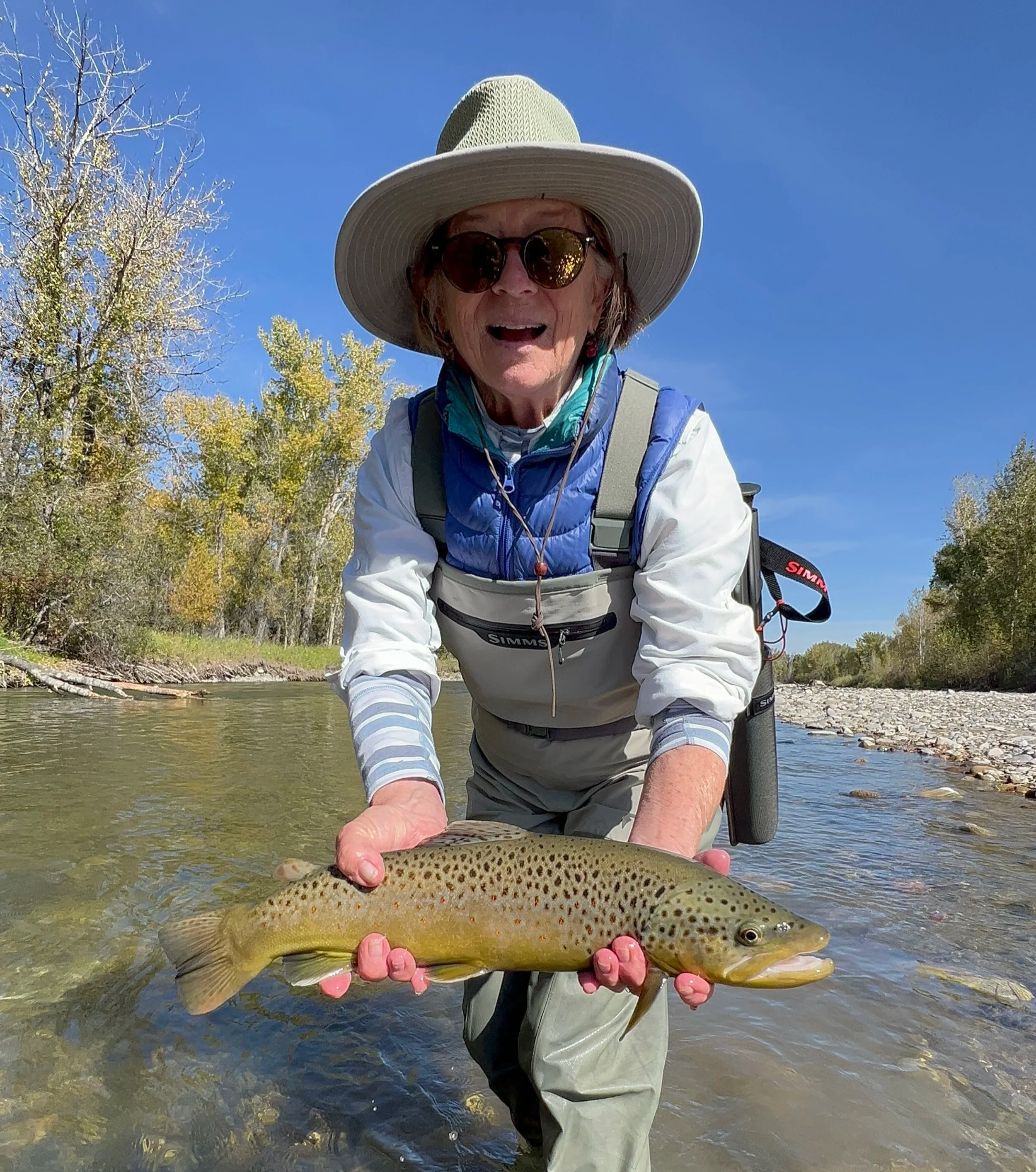 A angler on a fly fishing trip holding a large brown trout in a river with trees and clear sky in the background.