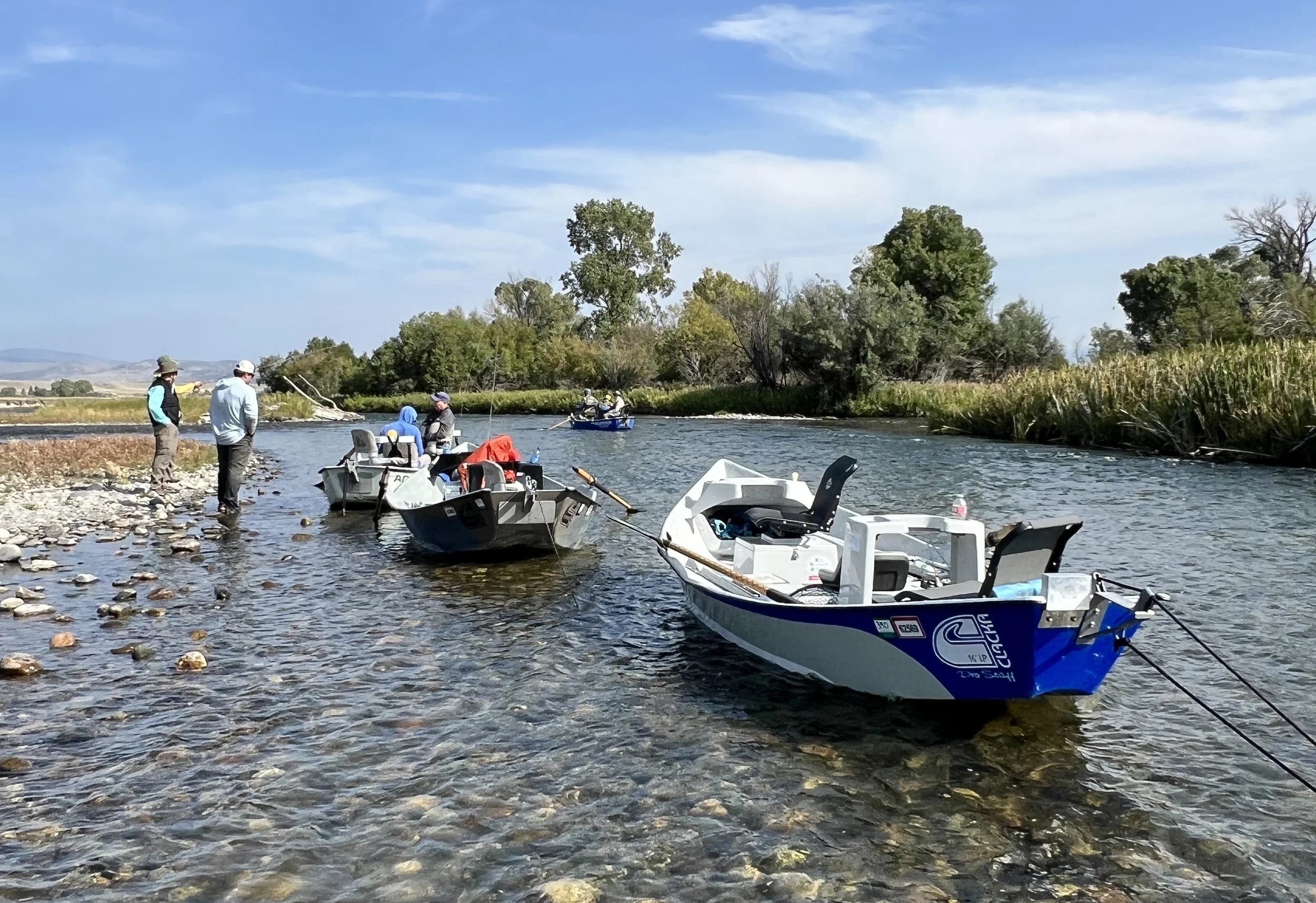 People launching drift boats into a river, with some standing on the rocky shore and others in the boats, surrounded by greenery and trees, under a partly cloudy sky.