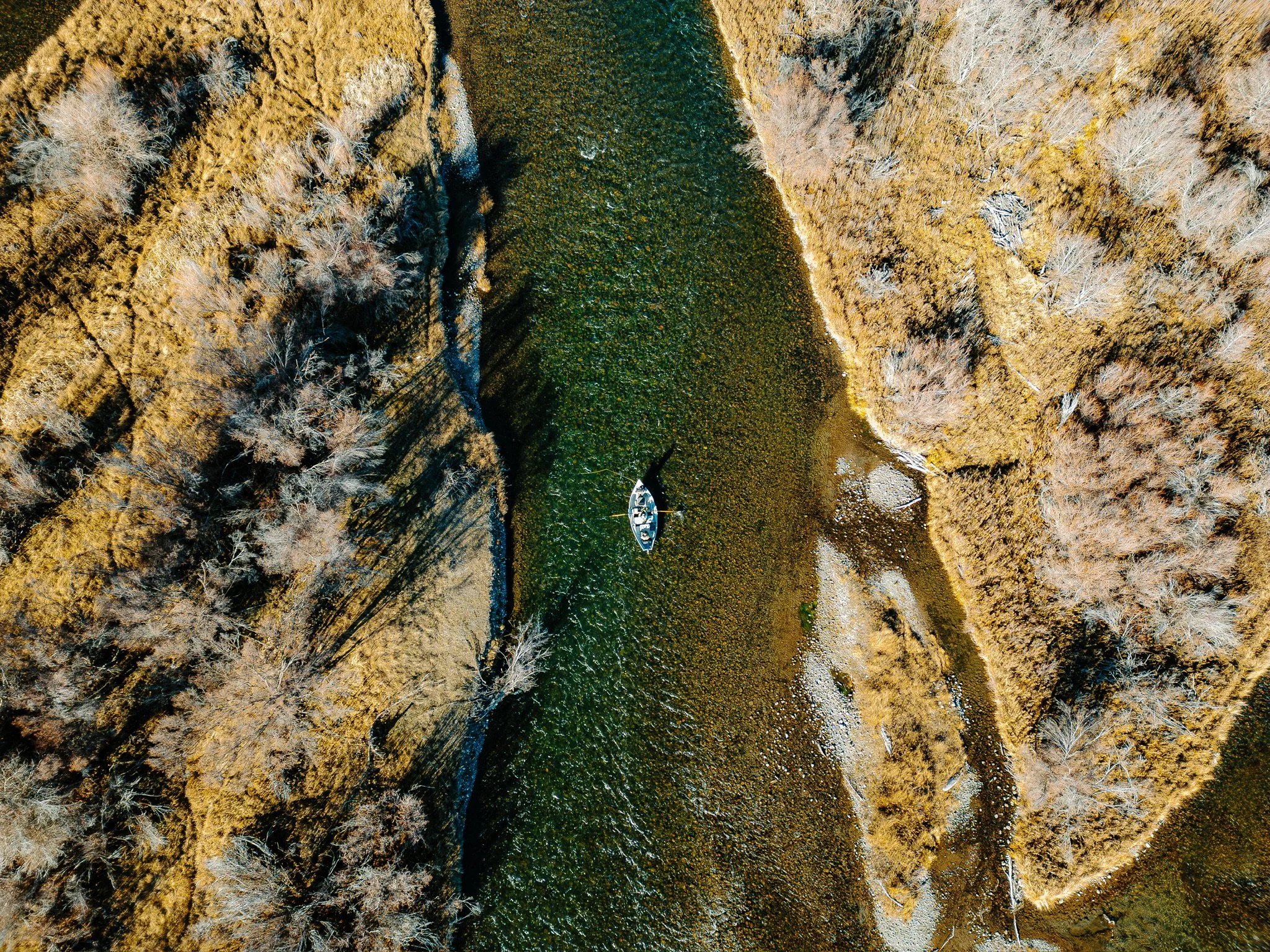 Aerial view of a drift boat on the Madison river flowing between trees and grassy areas during fall.