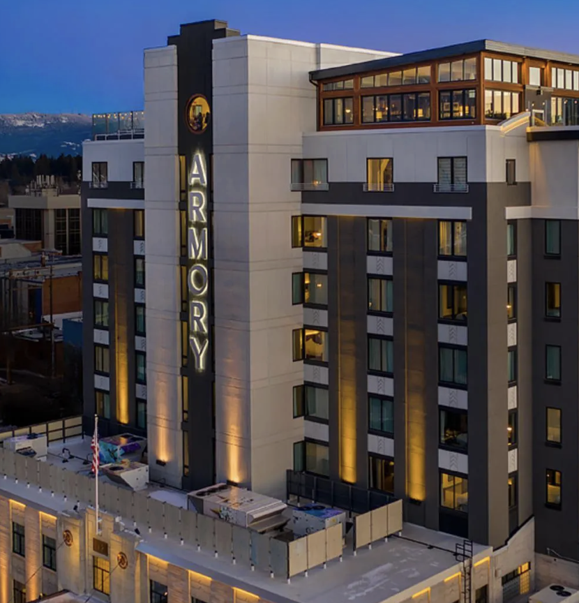 A modern multi-story hotel building labeled 'ARMORY' at dusk, with lit windows, balconies, and rooftop terrace, with mountains in the background.