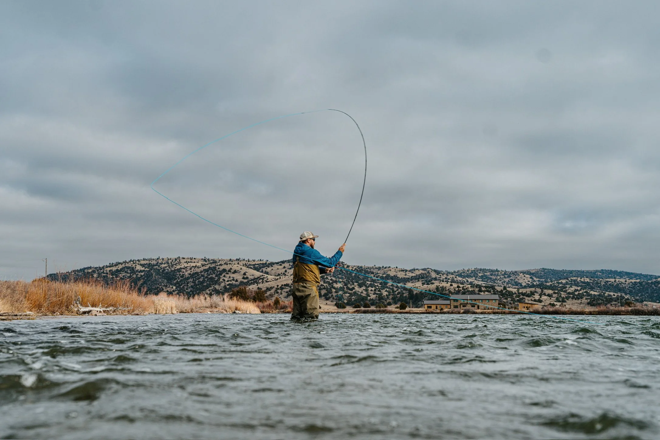 A man fly fishing in a river, with hills and cloudy sky in the background.