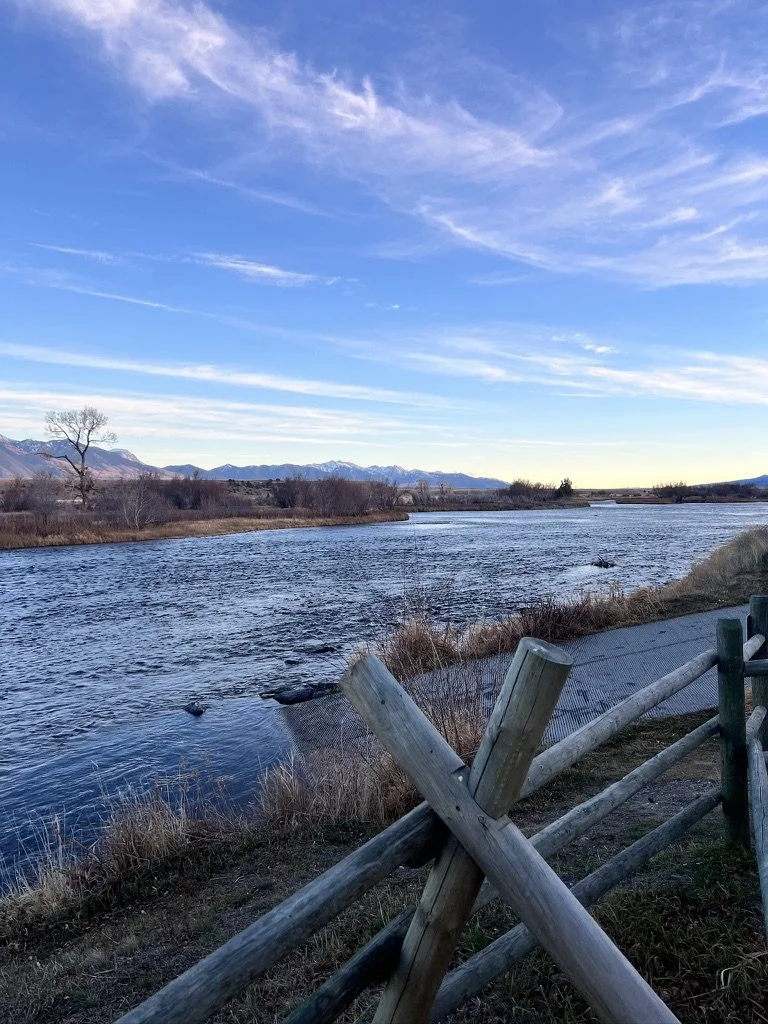 A scenic river landscape with mountain ranges in the background, a partly cloudy sky, leafless trees, dry grass, and a wooden fence in the foreground.