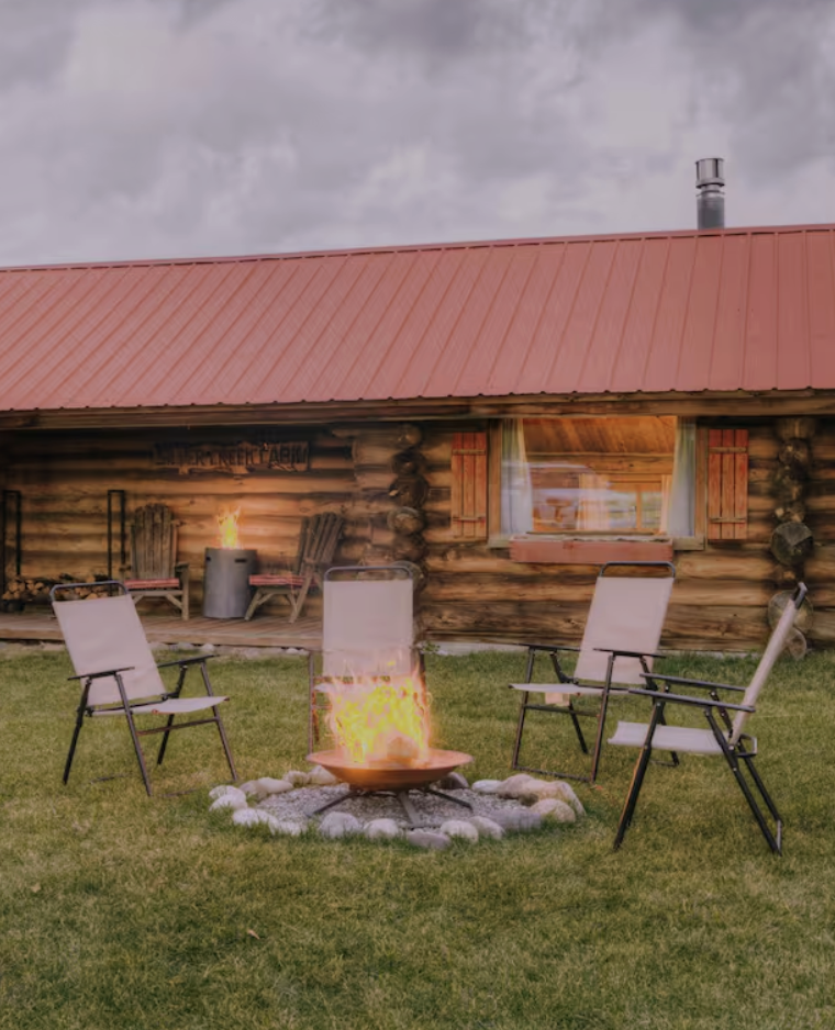 A cozy outdoor fire pit surrounded by four white camping chairs on a grassy lawn in front of a rustic log cabin with a red metal roof.