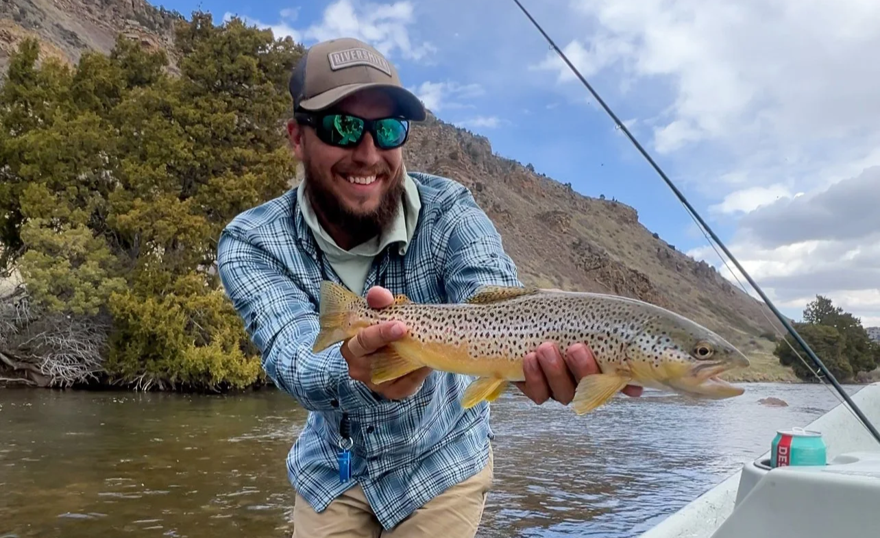 Man on a boat holding a freshly caught rainbow trout near a river with hills and trees in the background, on a partly cloudy day.