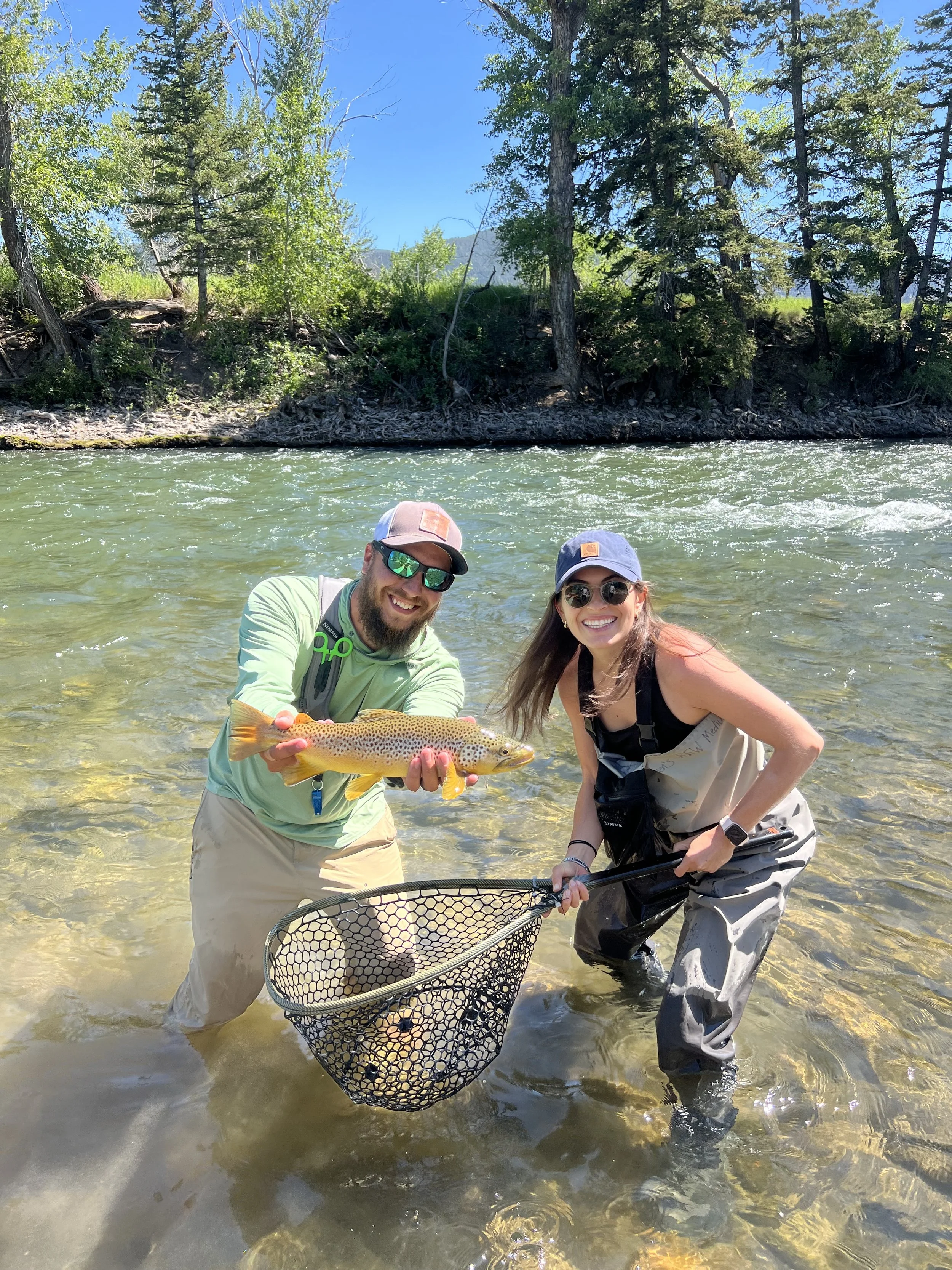 Smiling fishing guide and client standing in the gallatin river, holding a large fish, with a fishing net in front of them; trees and blue sky in the background.