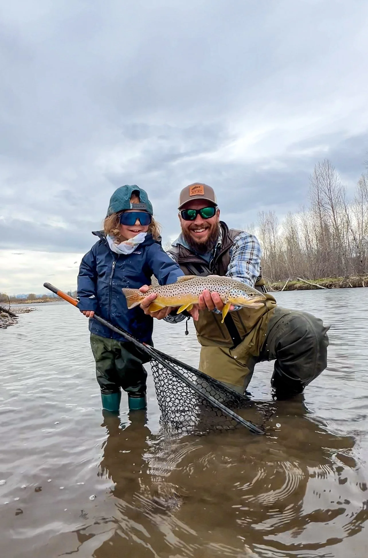A man and a young child in a river holding a large fish together, smiling and posing for the camera, with a cloudy sky and leafless trees in the background.