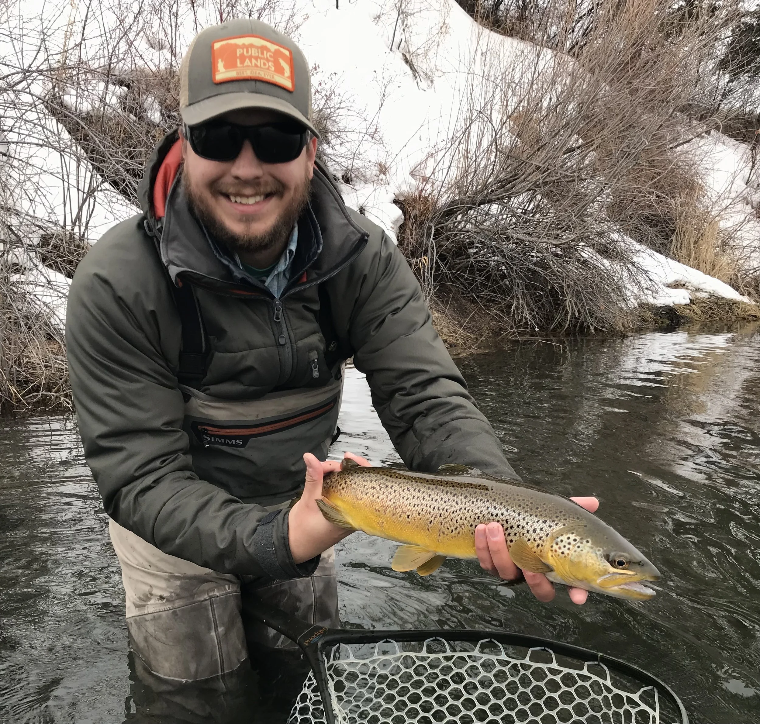 Man in outdoor gear smiling while holding a large rainbow trout fish near a riverbank with snow and bare bushes in the background.