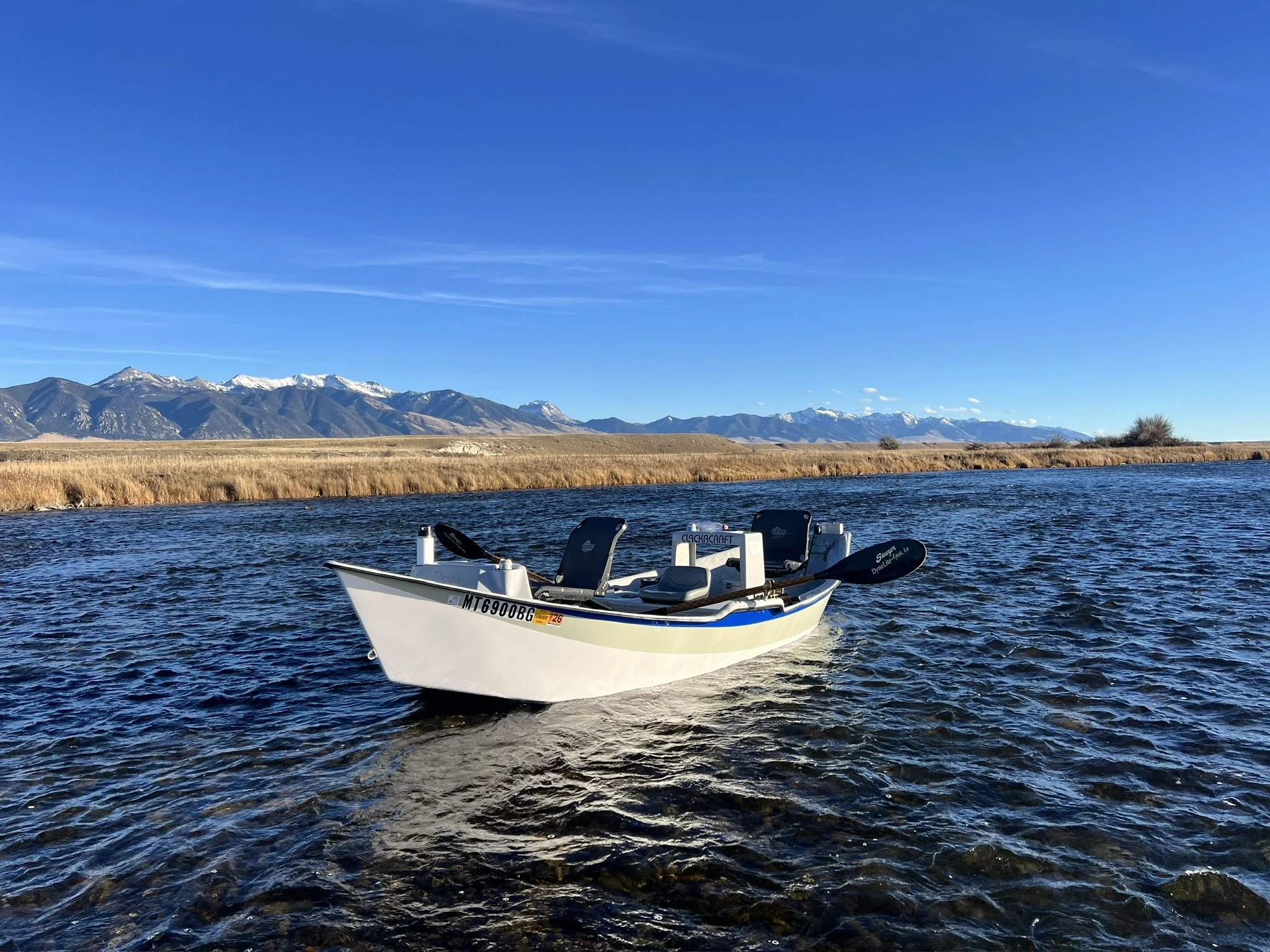 A Clackacraft Drift boat floating on the madison river with mountains in the background under a clear blue sky.