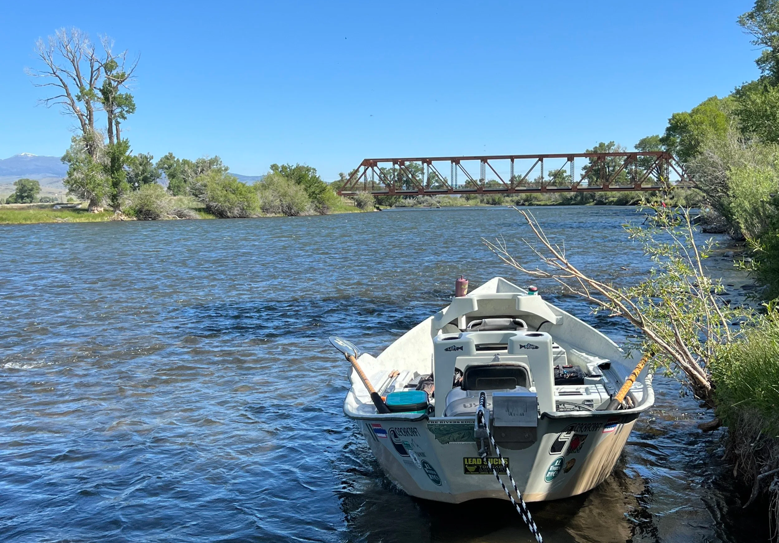 A white drift  boat tied to the shore of a river with green trees and bushes on either side, Varney bridge on the Madison River in the background, and mountains in the distance under a clear blue sky.