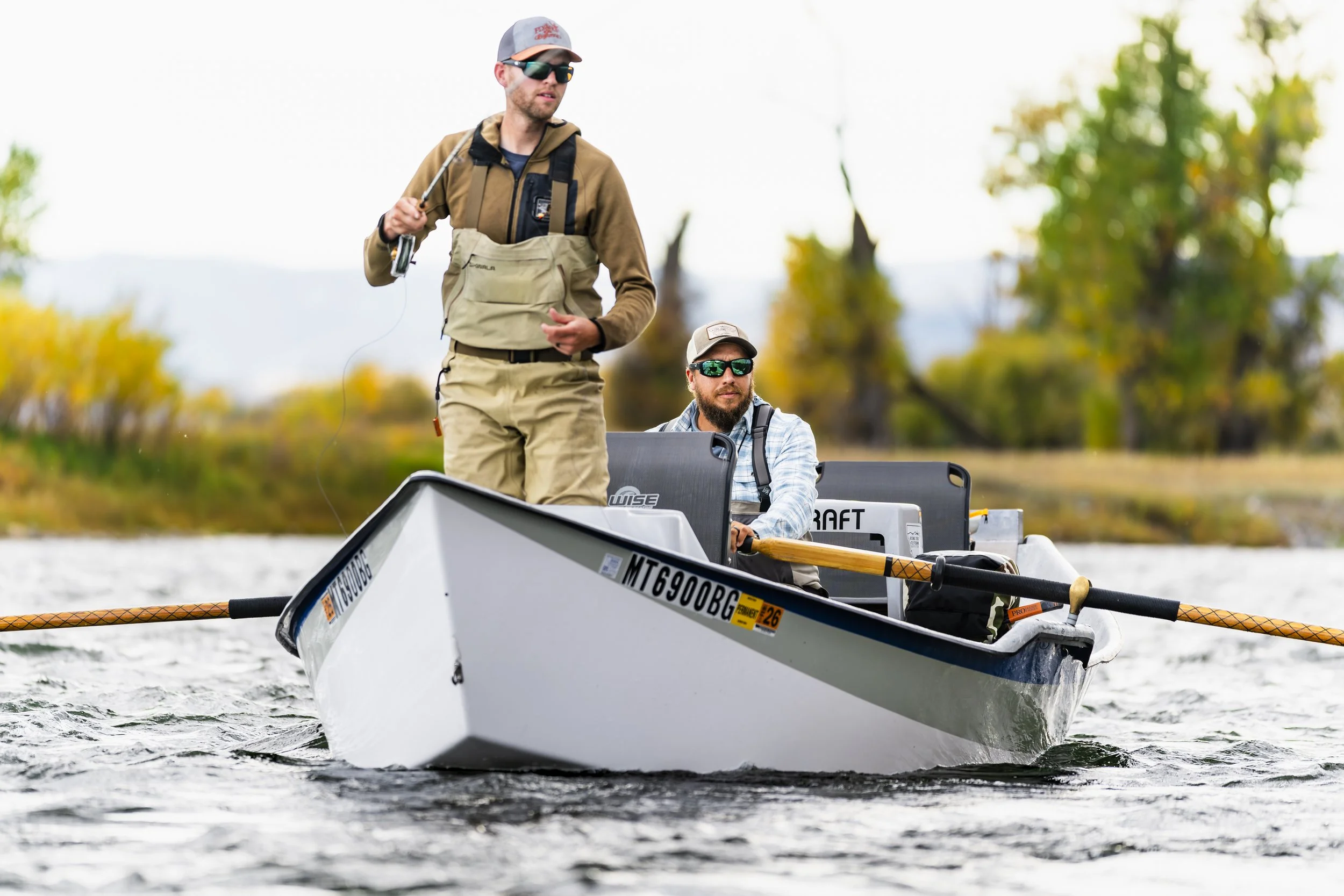 Two men fishing on a drfit boat on the Madison River in Fall with trees and cloudy sky in the background.
