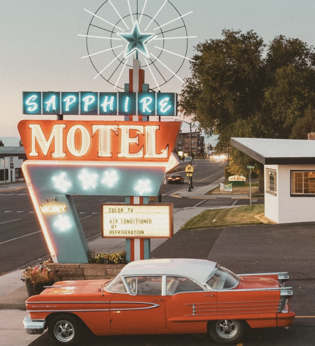 A vintage red and white car parked in front of the Sapphire Motel neon sign at dusk, with a small building to the right, trees, and a street with cars in the background.