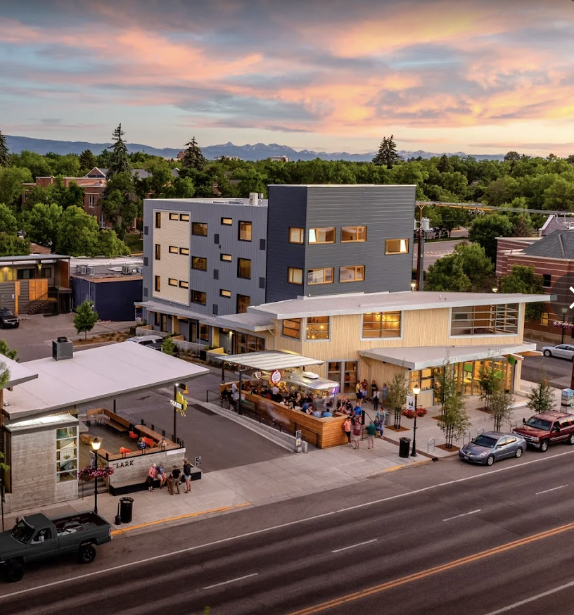 A modern multi-story building with a gray and beige exterior, large windows, and a flat roof, located on a city street at sunset. There's an outdoor area with people gathered around a food truck and seating, with trees and mountains in the background.