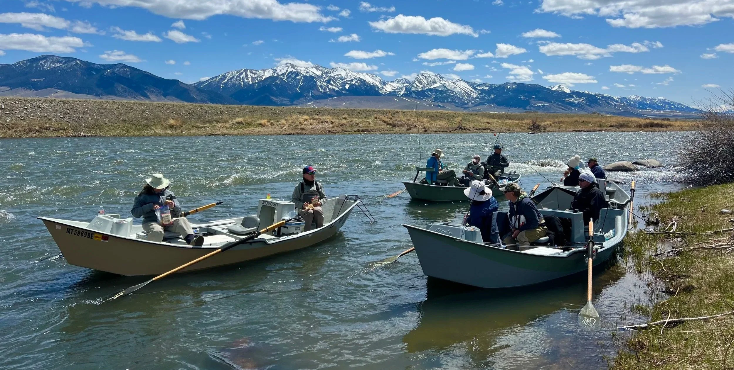 3 boats anchored up during a guide trip in may on the upper madison river. clients eating lunch in the boats with the madison mountain range snow covered in the background