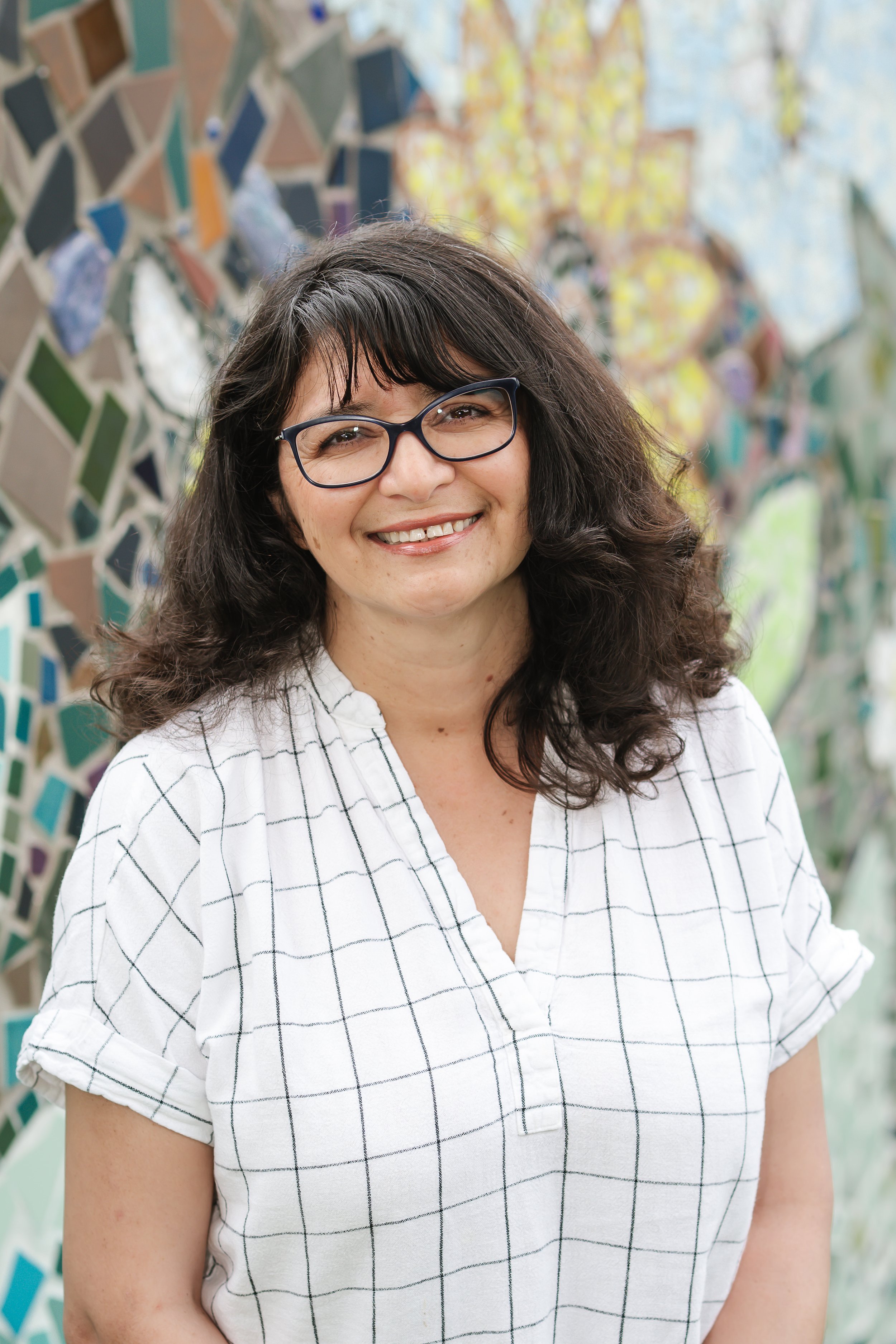 Smiling woman with dark hair, glasses, and a white shirt with black grid lines, standing in front of a colorful tile mosaic wall.
