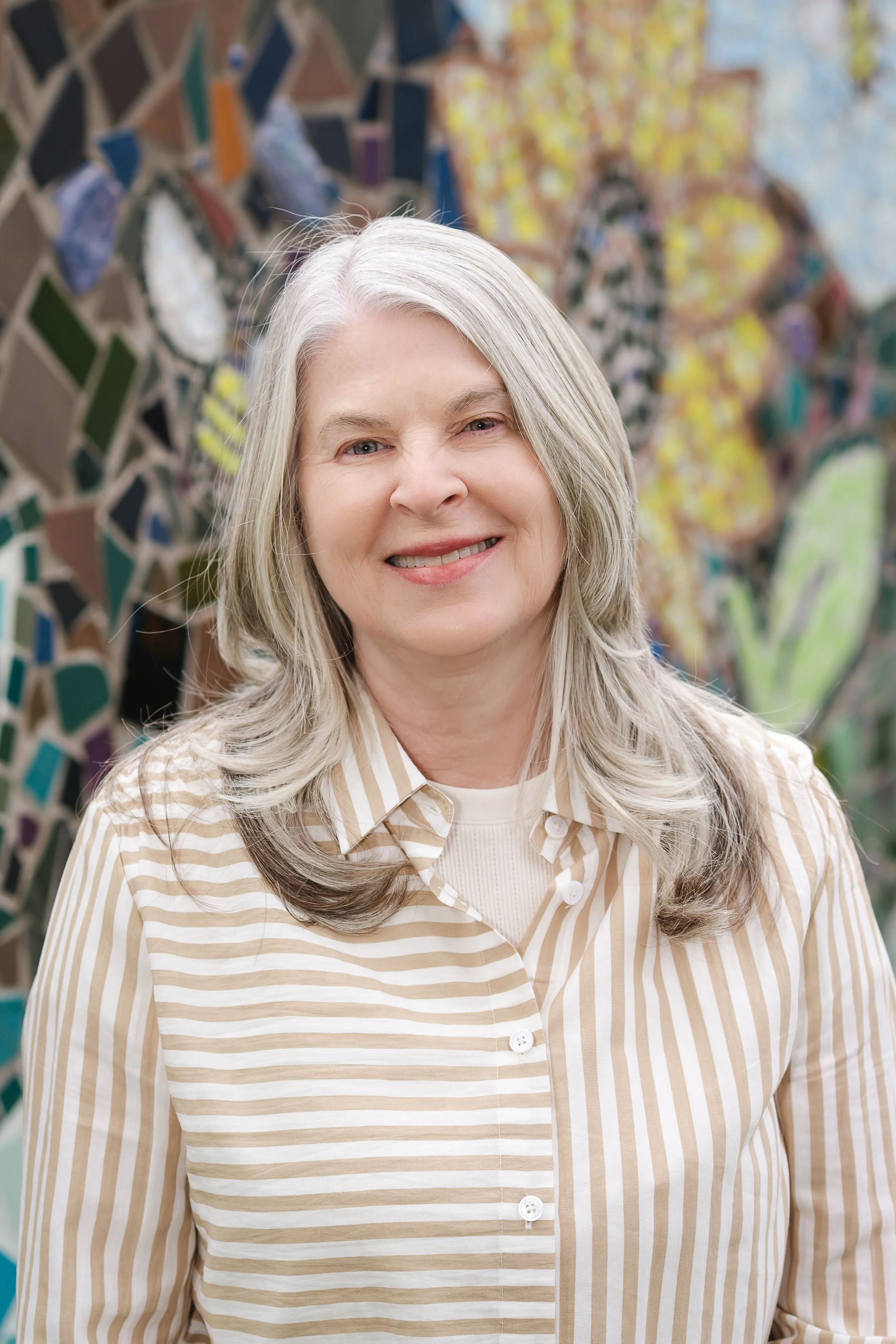 A woman with long gray hair smiling in front of a colorful mosaic wall.