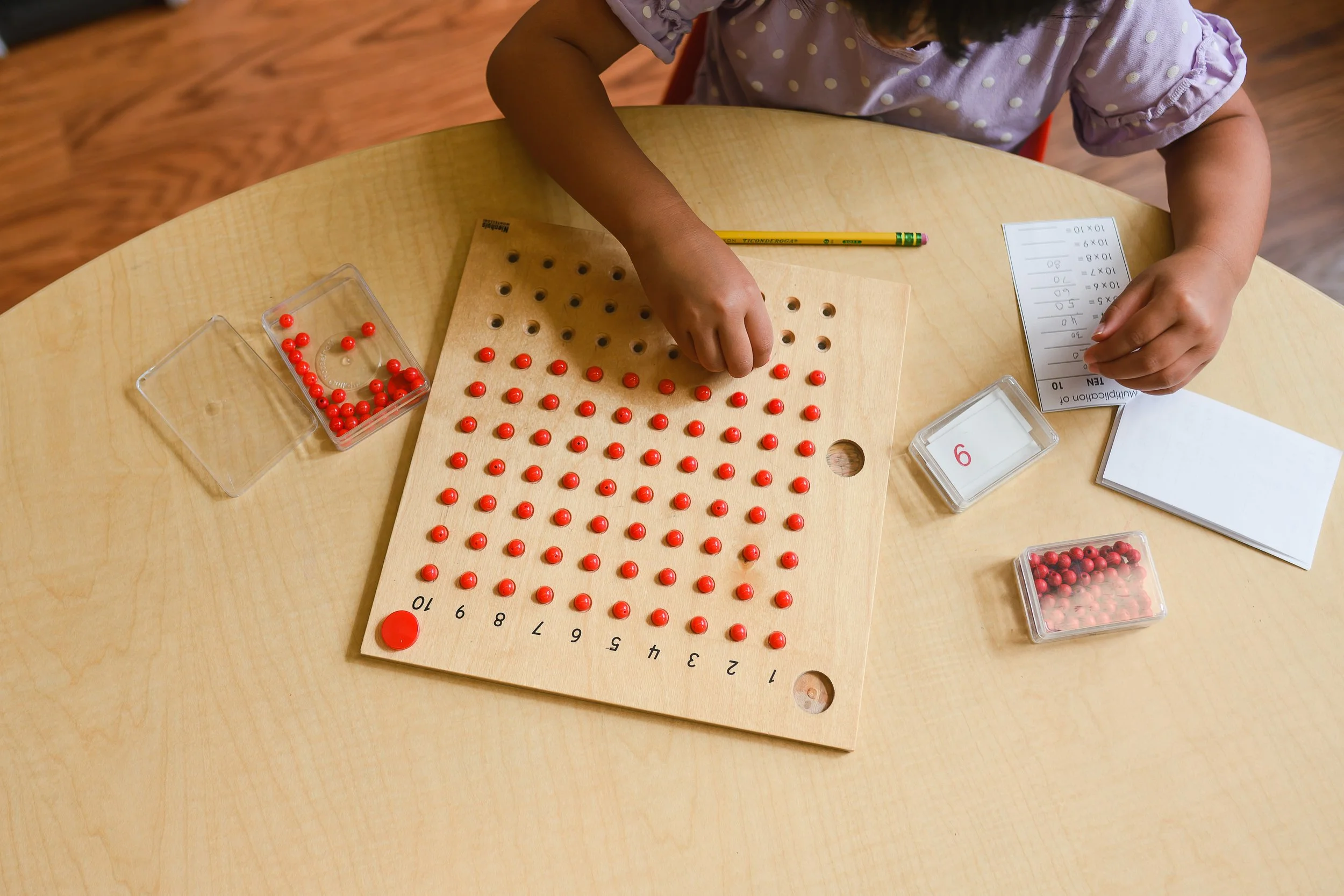 A child working on a math activity at a table, placing red counters on a math board, with a number chart, a pencil, and small plastic containers with red and pink counters nearby.