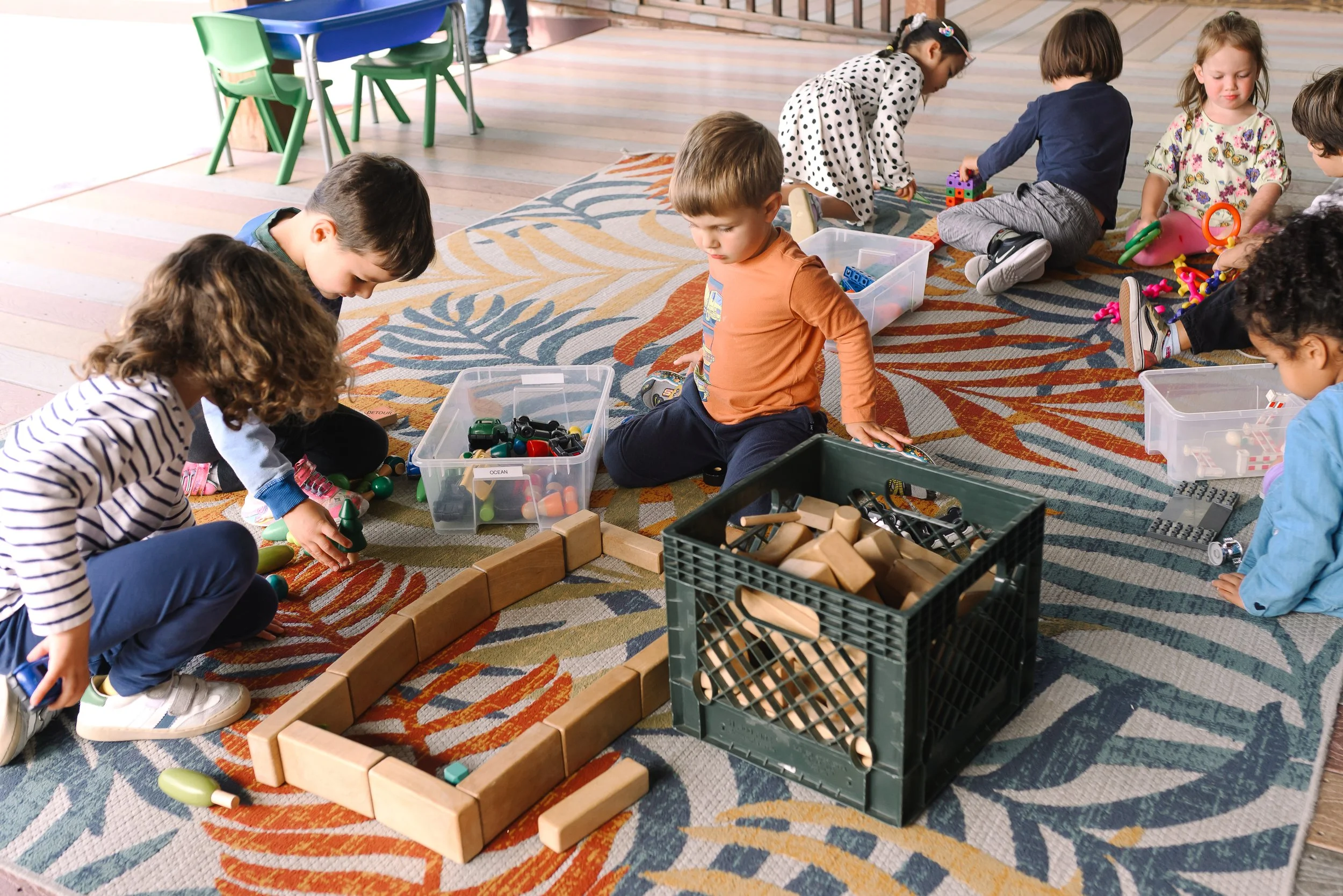 Children playing with toys on a colorful rug in a daycare or classroom setting, with tables and chairs in the background.