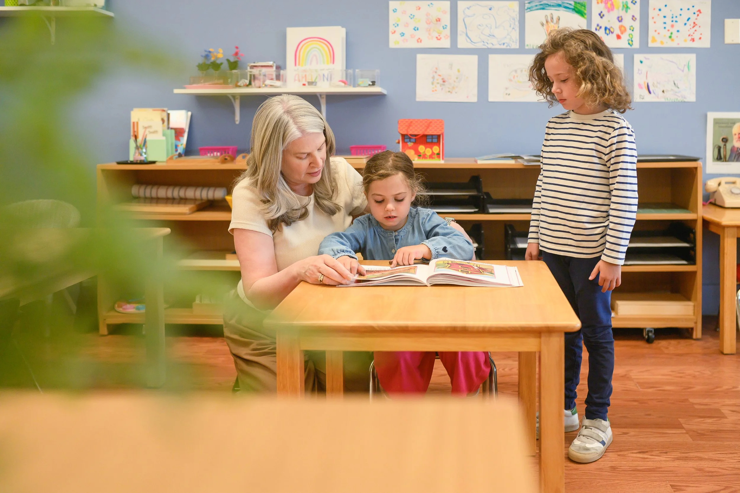 A woman and two young girls in a classroom looking at a picture book together. The classroom has colorful children's artwork on the walls and shelves with educational supplies.