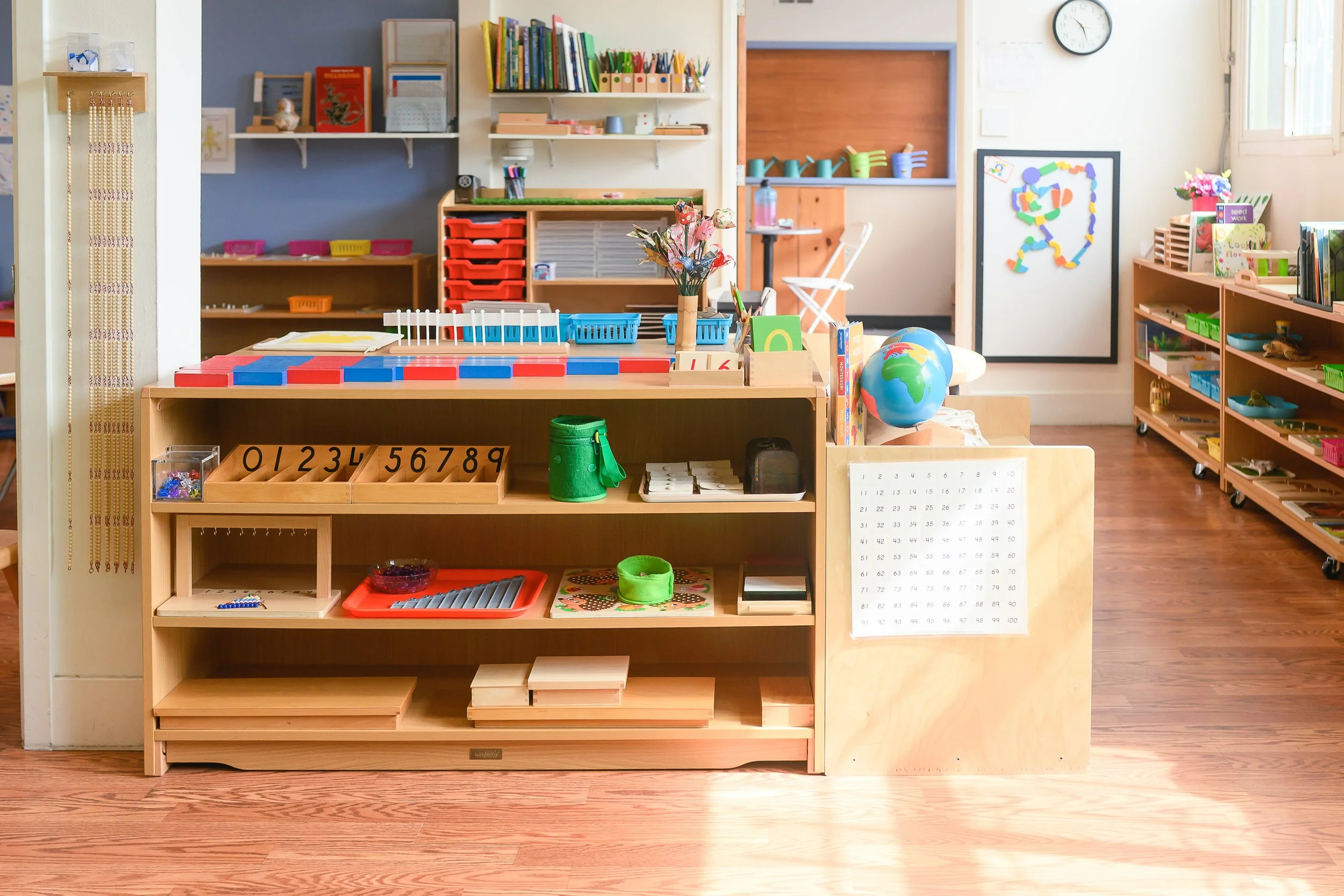 A classroom with shelves filled with educational materials, toys, and art supplies. There are tables and chairs suitable for young children, a globe, a clock on the wall, and colorful decorations.